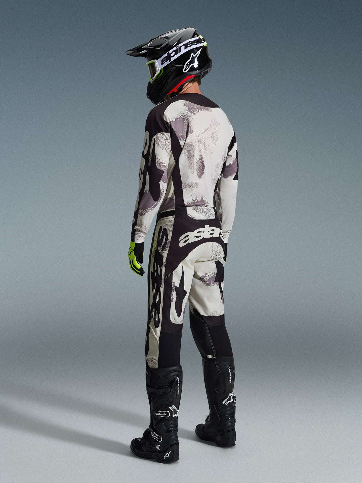 Conjunto de equipamento Alpinestars 2026 Racer Lahnd, piloto de motocross com camisola e calças a condizer em Sand Camo, padrão de camuflagem cinzento e preto com logótipos Alpinestars proeminentes, capacete preto, botas pretas e luvas amarelo néon, vista de perfil traseiro.