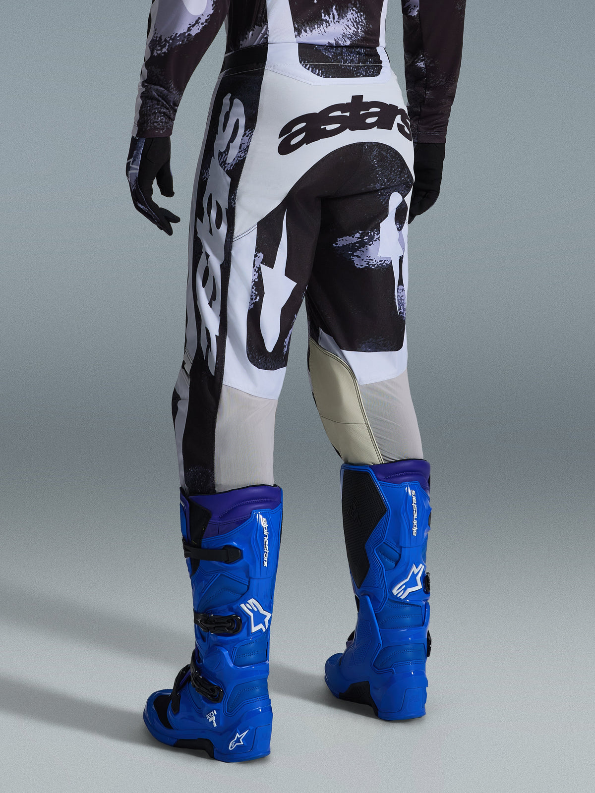 Alpinestars 2026 Racer Lahnd Pants, calças de motocross, padrão camuflado Iron Camo em cinzento e preto, vista traseira com grande logótipo astars a branco e preto, combinadas com botas de motocross em azul brilhante, equipamento profissional de corrida para todo-o-terreno