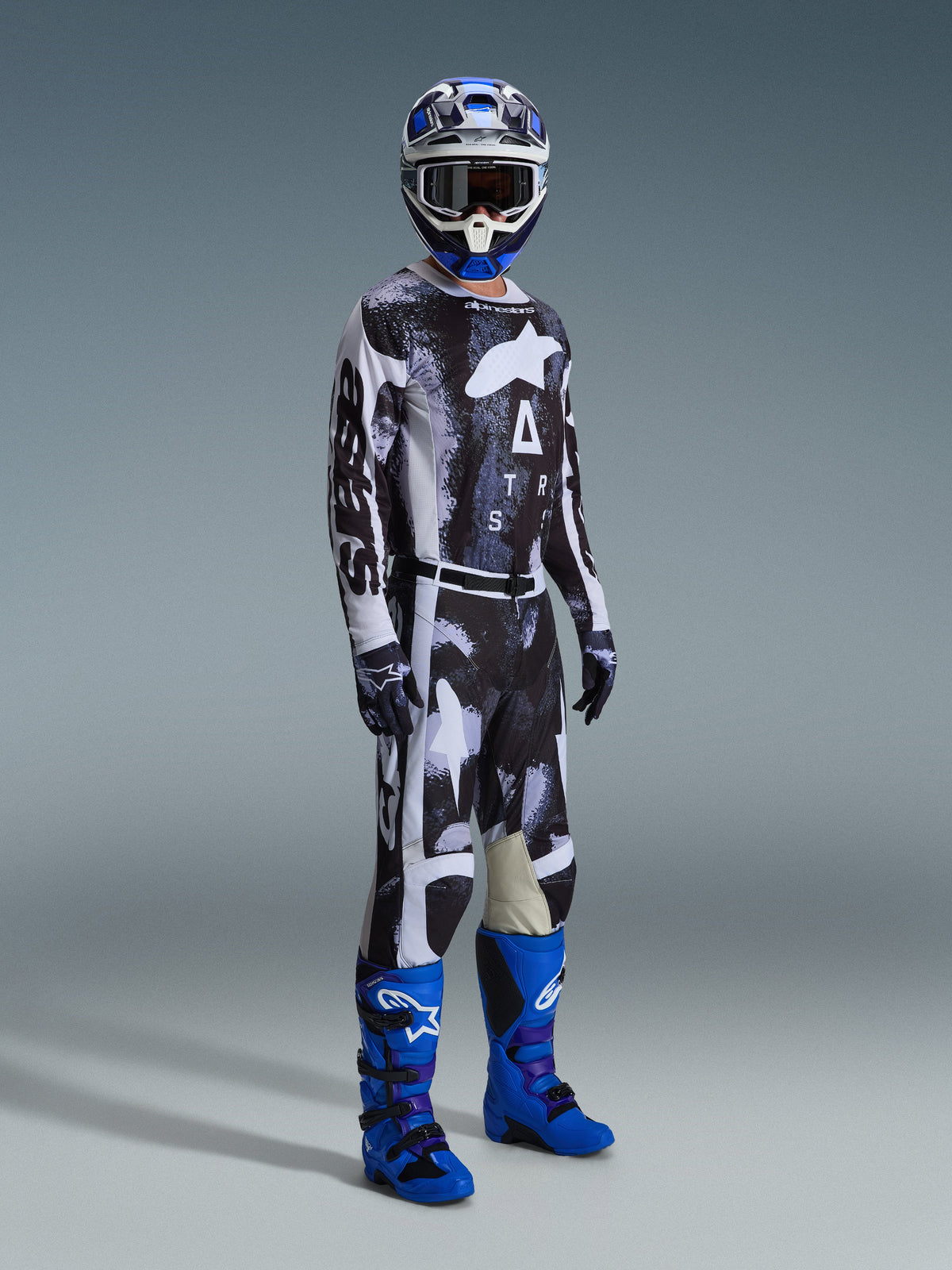 Piloto de motocross a usar a camisola Alpinestars 2026 Racer Lahnd e as calças a condizer em Iron Camo, com um padrão gráfico preto e branco, combinadas com botas azuis, um capacete azul e branco e luvas pretas, equipamento completo de corrida para todo-o-terreno