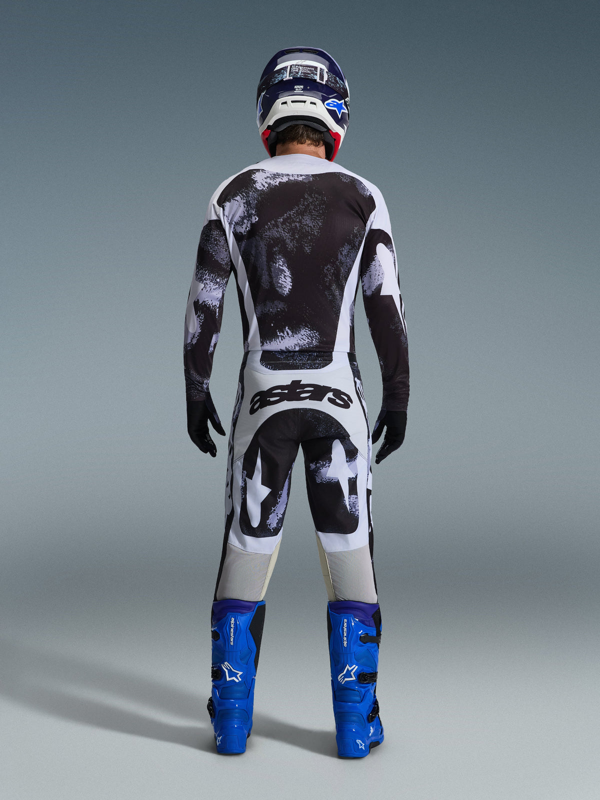 Piloto de motocross visto de costas a usar a camisola Alpinestars 2026 Racer Lahnd e calças a condizer em Iron Camo, com um padrão de salpicos em preto e branco, botas azul vivo e capacete coordenado, conjunto completo de equipamento para corridas off-road
