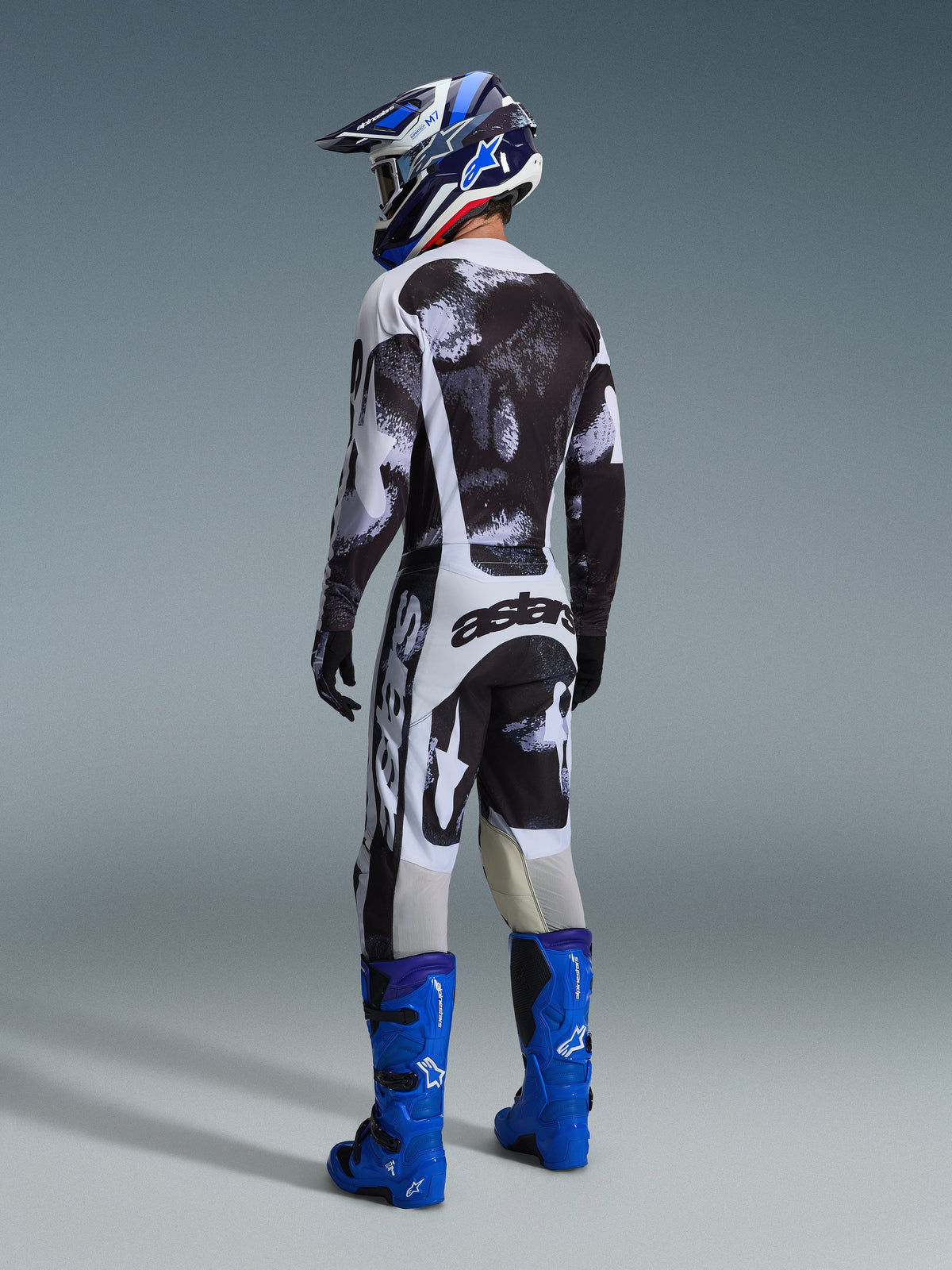 Piloto de motocross visto de costas com equipamento de corrida completo, camisola e calças Racer Lahnd 2026 em Iron Camo, padrão abstrato em preto, cinzento e branco, botas Alpinestars em azul brilhante, capacete azul e branco, conjunto coordenado para corridas todo-o-terreno