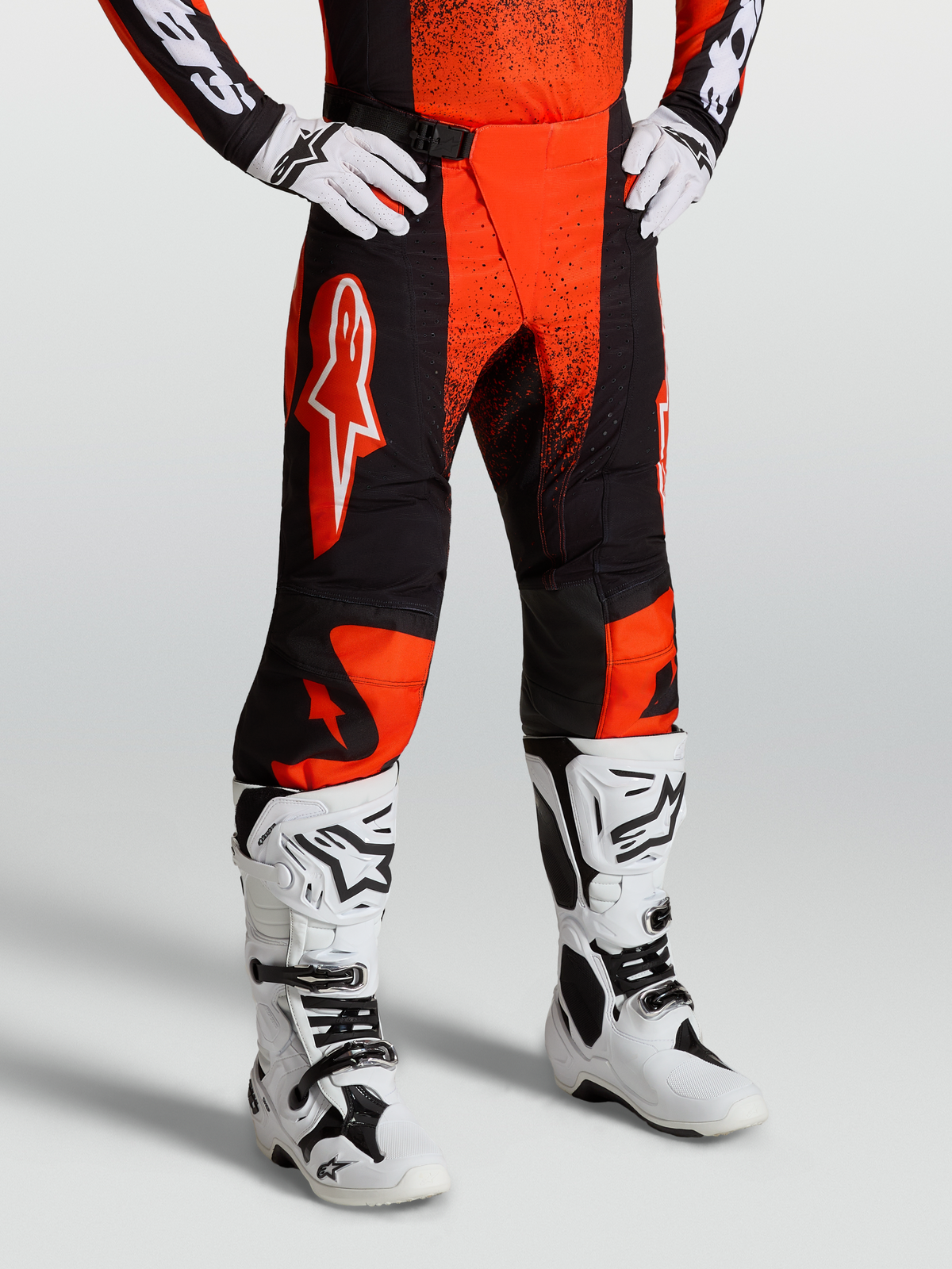 Alpinestars Calças Techstar Swan, calças de motocross para homem, vermelhas e pretas, com painéis elásticos e grafismos brancos, vestidas com botas de moto brancas.