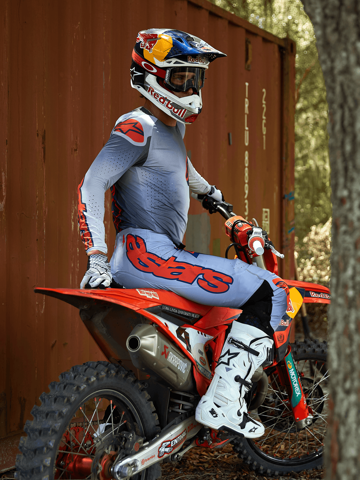 Piloto de motocross sentado numa mota de motocross vermelha, a usar o conjunto de equipamento Alpinestars 2025 Supertech Lipan em cinzento e laranja fluorescente, calças azul-claro com logótipo Astars laranja em destaque, camisola a condizer, botas de motocross brancas e capacete Red Bull, equipamento de corrida profissional coordenado