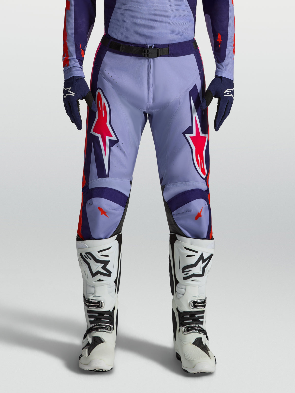 Alpinestars calças Racer Air Hunt, calças de motocross em violeta claro e roxo profundo com detalhes rosa fluorescente, design ventilado, usadas com botas brancas.