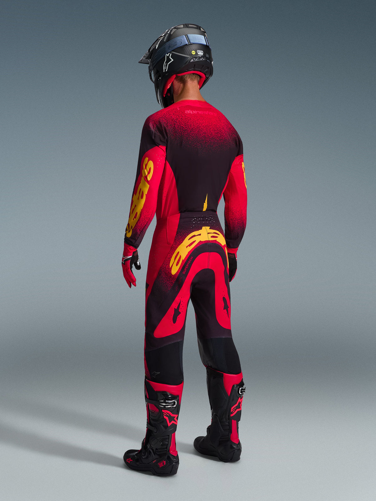Piloto de motocross visto de costas a usar calças Alpinestars 2026 Supertech Scenz em vermelho, preto e amarelo, camisola a condizer com marca amarela em destaque, botas de motocross pretas e vermelhas e capacete preto, conjunto completo de equipamento de competição para performance em todo-o-terreno