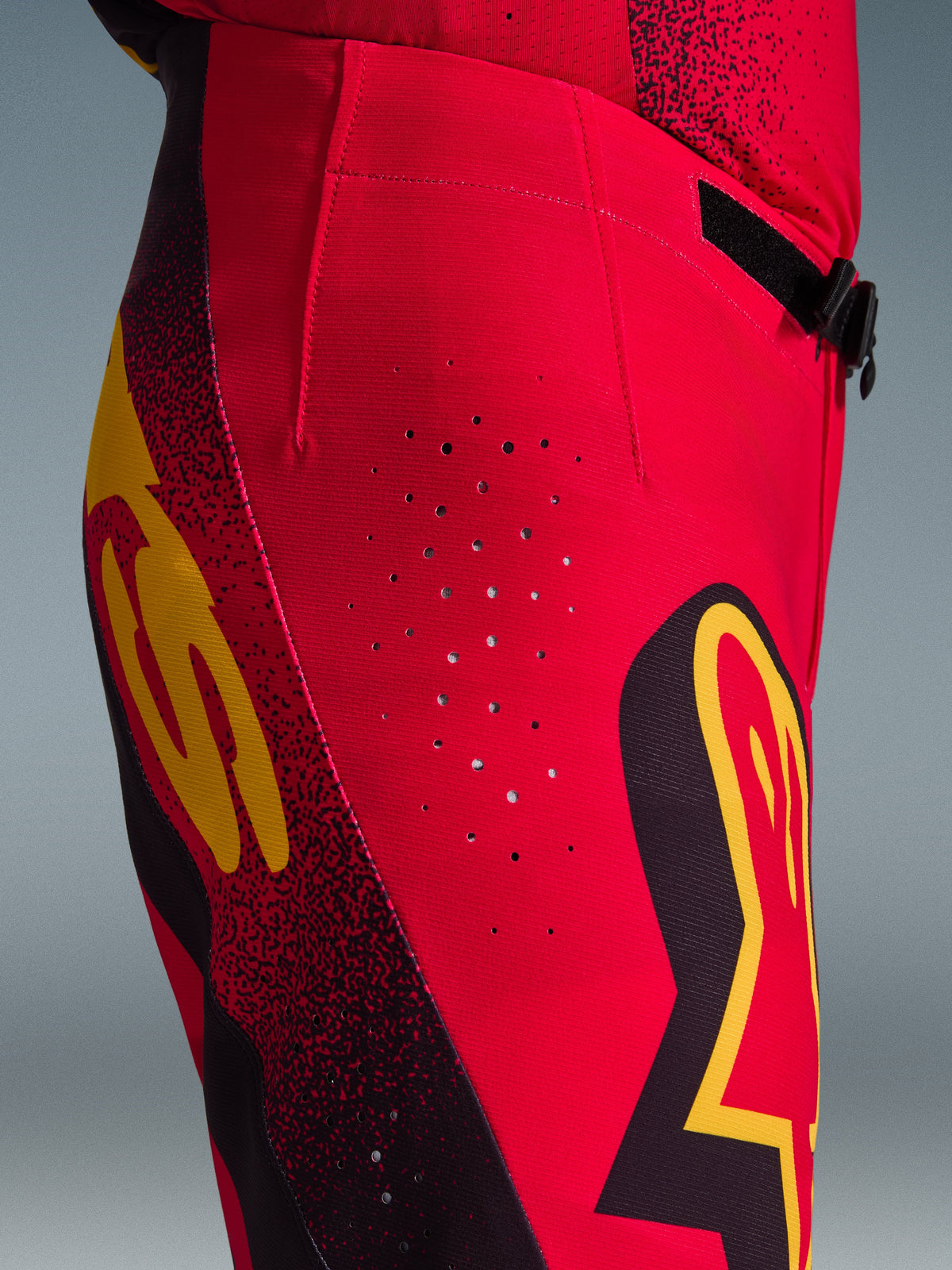 Alpinestars 2026 Supertech Scenz Pants, calças de motocross, vermelhas, pretas e amarelas, vista lateral em pormenor com perfurações de ventilação cortadas a laser, logótipos gráficos arrojados e tecido rip-stop elástico de alto desempenho para corridas de motocross.