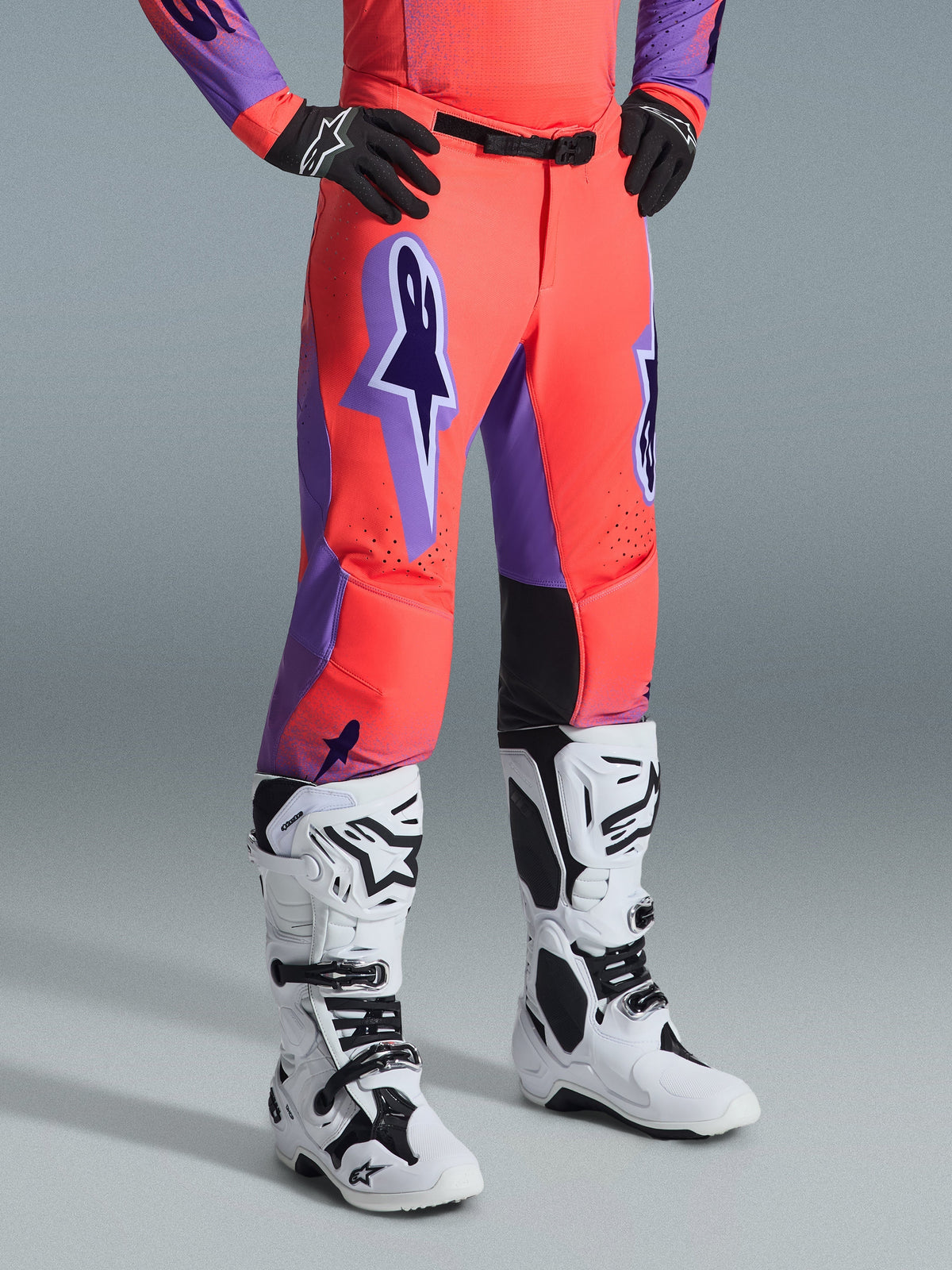 Alpinestars 2026 Supertech Scenz Pants, calças de motocross, Hot Coral e roxo, usadas com botas de motocross brancas e pretas e luvas pretas, com um logótipo Alpinestars roxo grande, painéis internos dos joelhos em couro preto e design com ajuste atlético de corrida