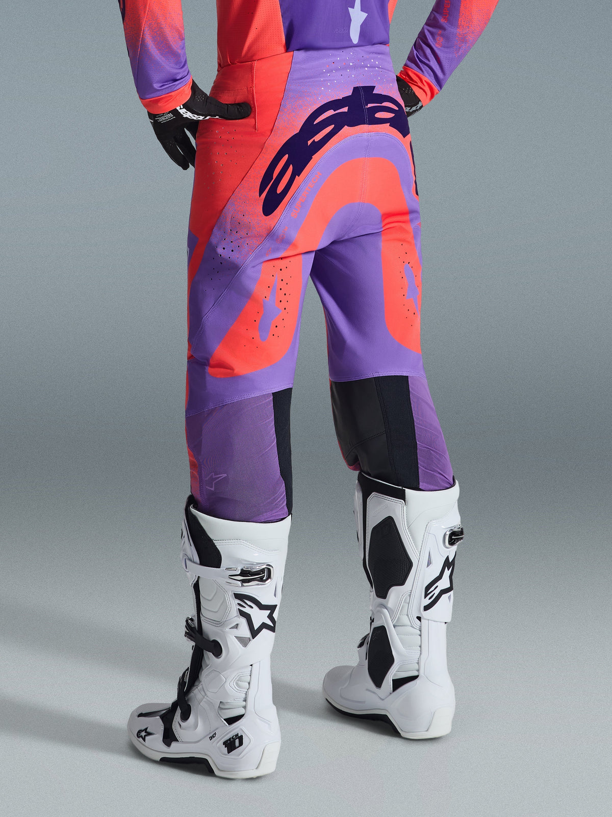 Alpinestars 2026 Supertech Scenz Pants, calças de motocross, coral vibrante e roxo, vista traseira com branding arrojado no assento, painéis vibrantes em coral e roxo com zonas perfuradas para fluxo de ar, usadas com botas de competição brancas e pretas