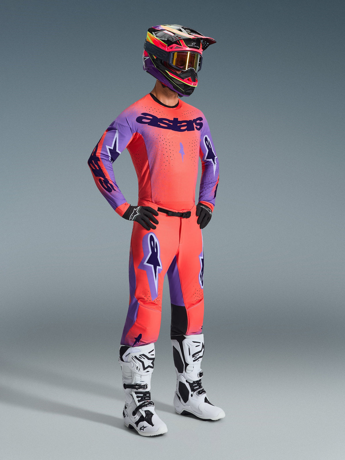 Piloto de motocross com equipamento completo de corrida, apresentando as calças Alpinestars 2026 Supertech Scenz em Hot Coral Purple, camisola em coral e roxo a condizer, capacete preto e rosa, botas brancas e pretas, kit de competição off-road profissional
