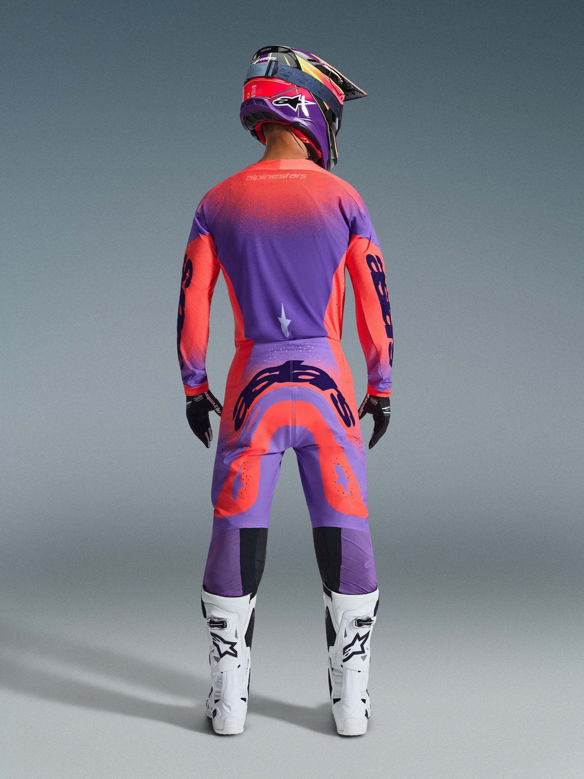 Alpinestars 2026 Supertech Scenz Pants, calças de motocross, Hot Coral e Roxo, vista traseira de um piloto de motocross a usar um conjunto completo de equipamento de corrida, com um design de gradiente vibrante e logótipos traseiros marcantes, combinado com camisola a condizer, capacete e botas brancas.
