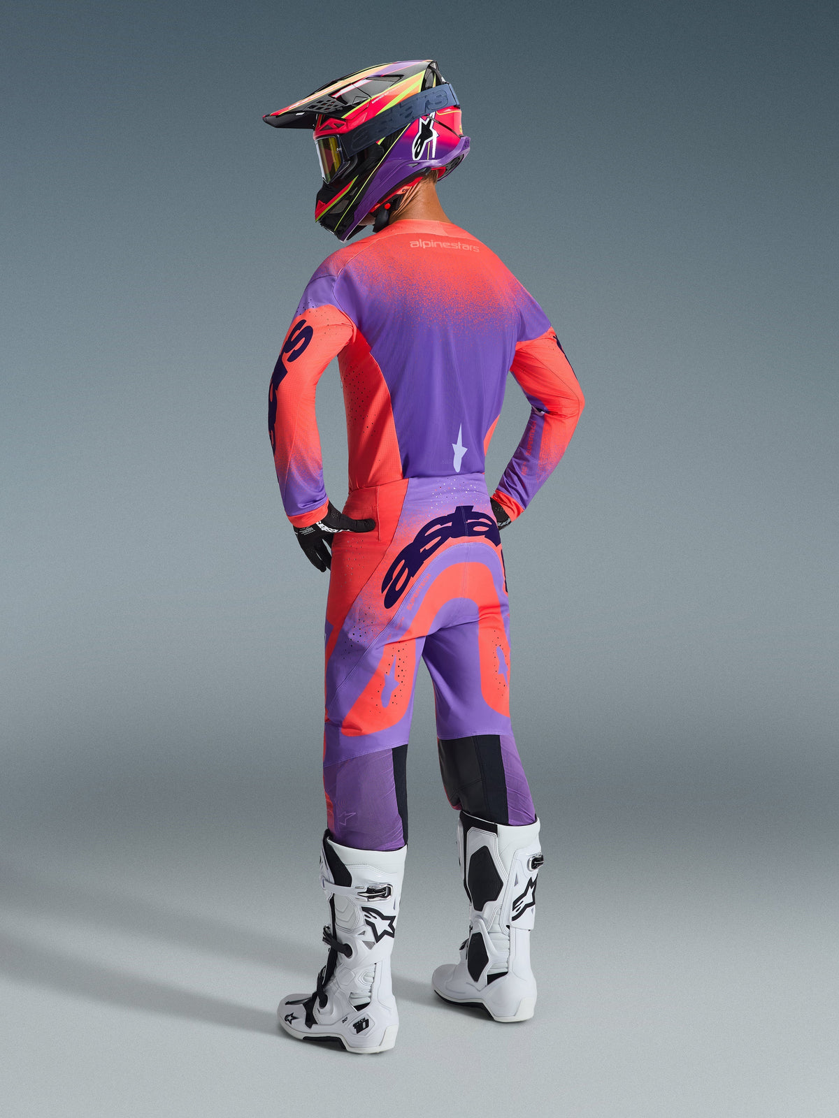Alpinestars 2026 Supertech Scenz Pants, calças de motocross, coral vibrante e roxo, vista traseira com logótipo marcante no assento, usadas por um piloto com camisola a condizer e botas brancas, equipamento profissional de corrida off-road