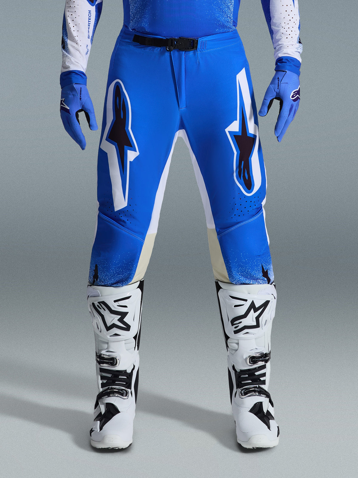 Alpinestars 2026 Supertech Scenz Pants, calças de motocross, azul e branco, usadas com luvas azuis a condizer e botas brancas, com grafismos grandes a branco e preto nas coxas, corte atlético para desempenho em off-road