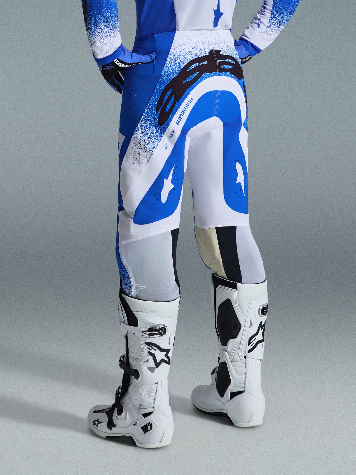 Calças Alpinestars Supertech Scenz 2026, calças de motocross, azul UCLA e branco, vista traseira mostrando o grande logótipo Astars preto no assento e a marca 'SUPERTECH', usadas com botas Tech 10 brancas e uma camisola azul e branca a condizer.
