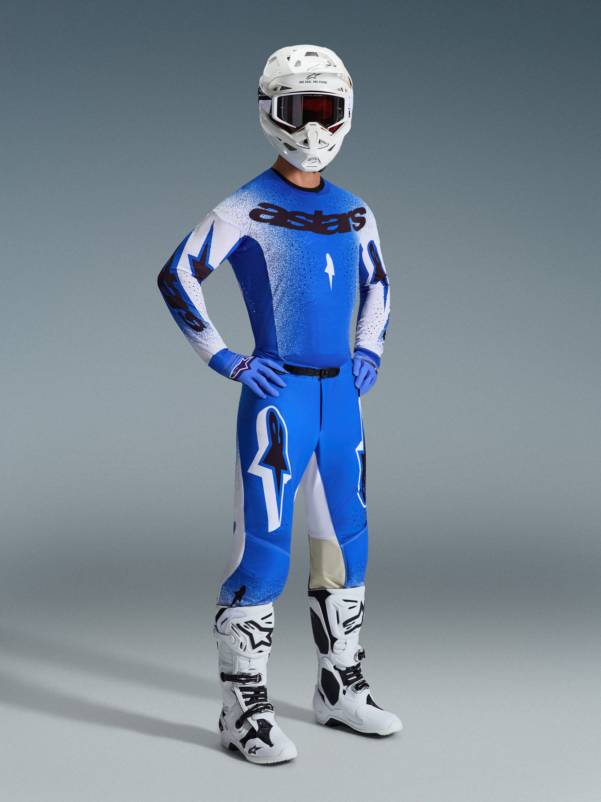 Piloto de motocross com equipamento de corrida completo, destacando as calças 2026 Supertech Scenz em Azul Ucla e Branco, camisola azul e branca com logótipo Astars, capacete branco com óculos vermelhos e botas brancas e pretas, conjunto coordenado para competição profissional de off-road