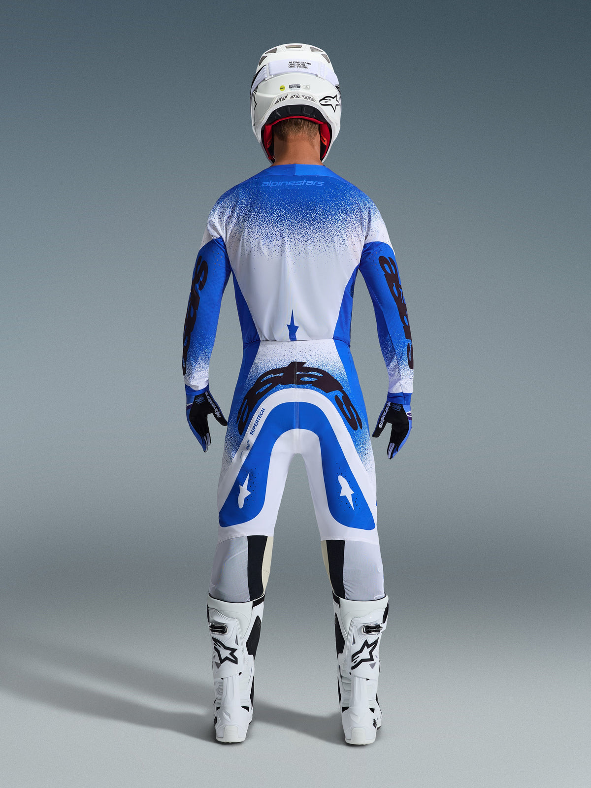 Piloto de motocross visto de trás com equipamento de corrida completo, calças Alpinestars 2026 Supertech Scenz em azul Ucla e branco, camisola azul e branca a condizer com padrão salpicado, capacete branco, botas brancas e pretas, conjunto coordenado para competição profissional
