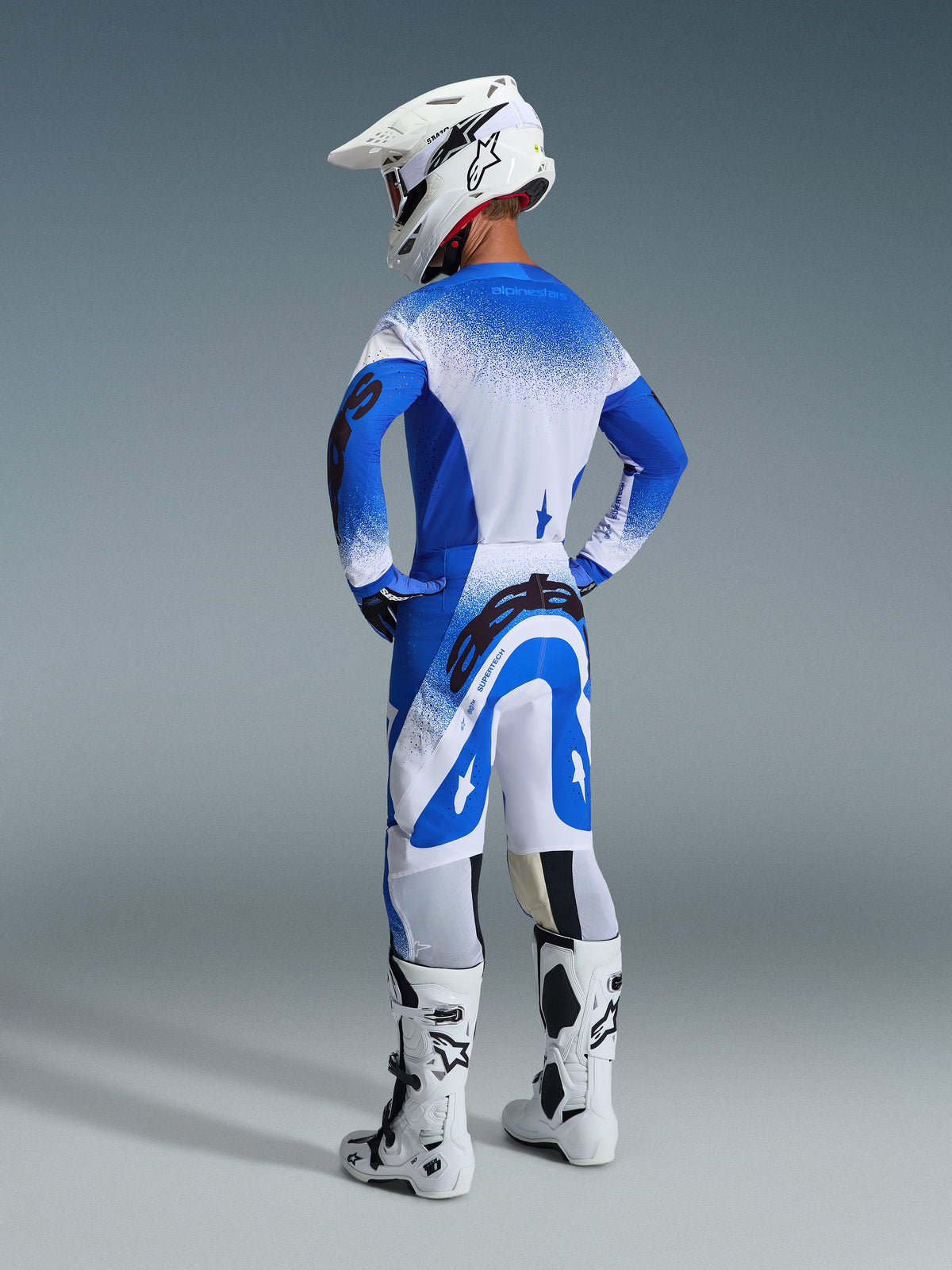 Piloto de motocross visto de costas a usar as calças e camisola 2026 Supertech Scenz em azul Ucla Blue e branco, capacete branco com logótipo Alpinestars, botas de motocross brancas e pretas, conjunto de equipamento profissional de corrida off-road.
