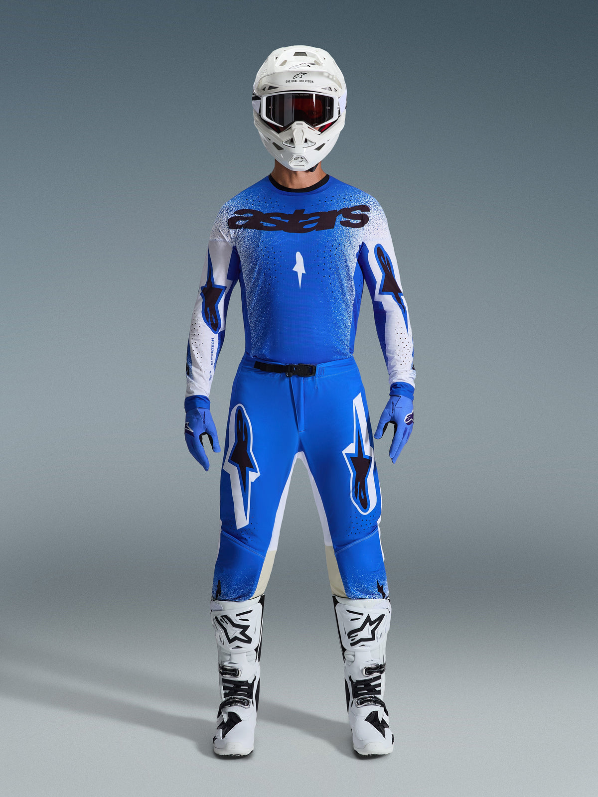 Piloto de motocross com equipamento de corrida completo, camisola azul e branca com marca astars a preto, Calças Supertech Scenz 2026 em azul e branco com logótipos de estrela destacados, capacete branco, luvas azuis e botas brancas, conjunto de corrida atlético coordenado