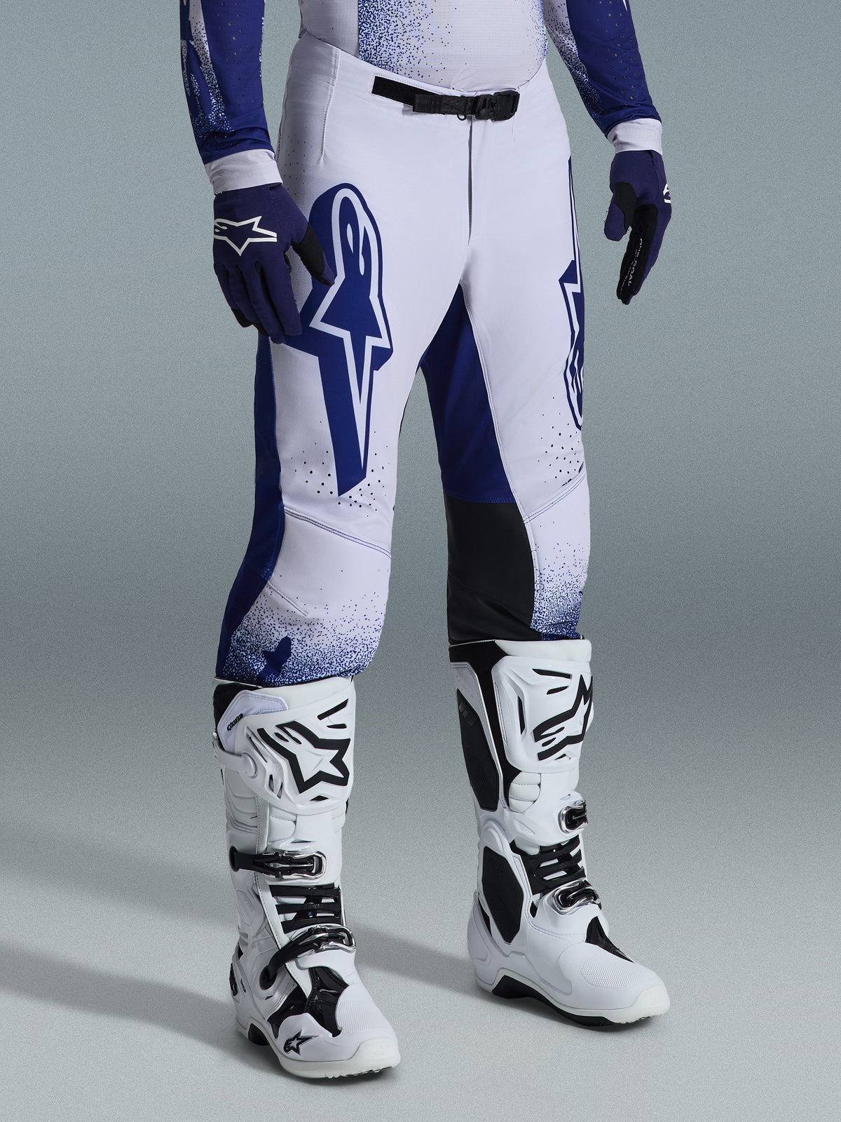 Calças Alpinestars 2026 Supertech Scenz, calças de motocross, cinzento claro e azul escuro, usadas com botas de motocross brancas e luvas azuis, com grandes logótipos Alpinestars azuis nas coxas, painéis pretos nos joelhos e um design gráfico pontilhado, equipamento profissional de corrida todo-o-terreno