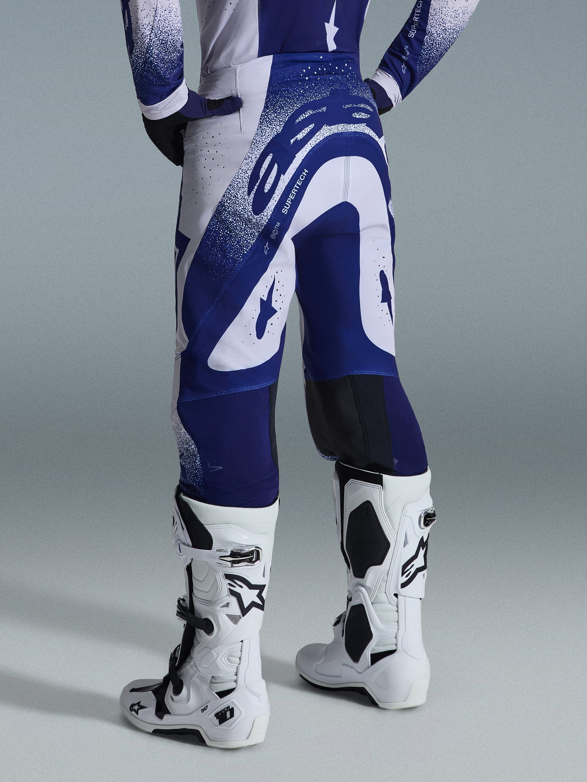 Alpinestars 2026 Supertech Scenz Pants, calças de motocross, cinzento claro e azul escuro, vista traseira, usadas com camisola a condizer e botas brancas, com grafismos azuis marcantes e detalhes salpicados, ajuste atlético de alta performance para corridas todo-o-terreno
