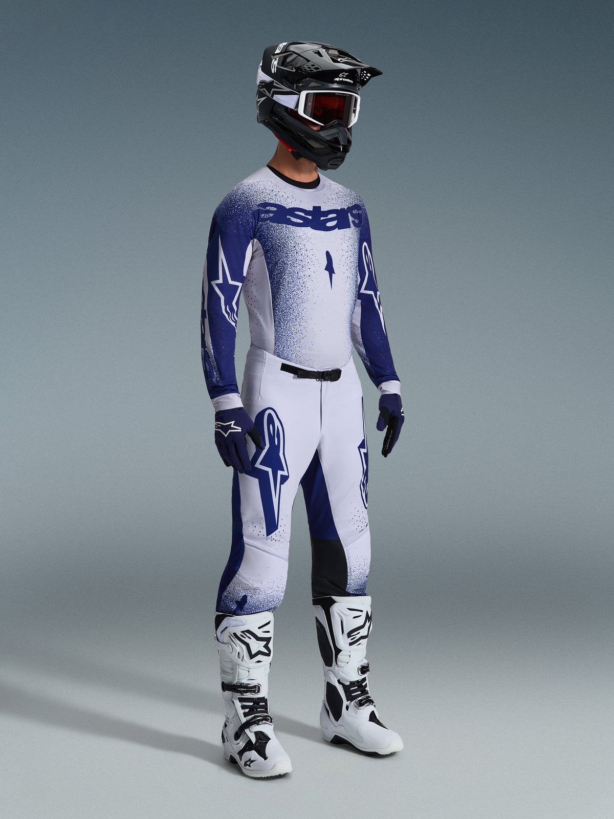Piloto de motocross vestido com as Calças Supertech Scenz 2026 em cinzento claro e azul escuro, camisola azul e branca a condizer com a marca 'astars' e efeito de pintura em spray, capacete preto com óculos de proteção brancos, botas de motocross brancas e pretas, equipamento profissional de corrida todo-o-terreno coordenado.
