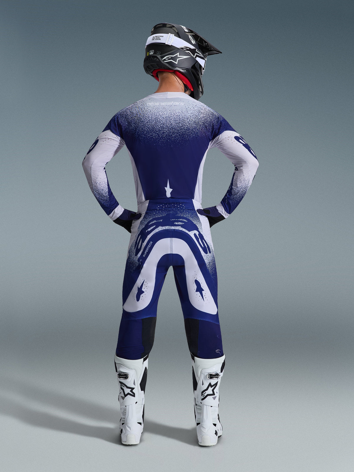 Alpinestars 2026 Supertech Scenz Pants, calças de motocross, cinzento claro e azul escuro, vista traseira de um piloto a usar um conjunto de equipamento a condizer com gráficos de gradiente salpicado, botas brancas e capacete preto, ajuste atlético profissional para corridas todo-o-terreno