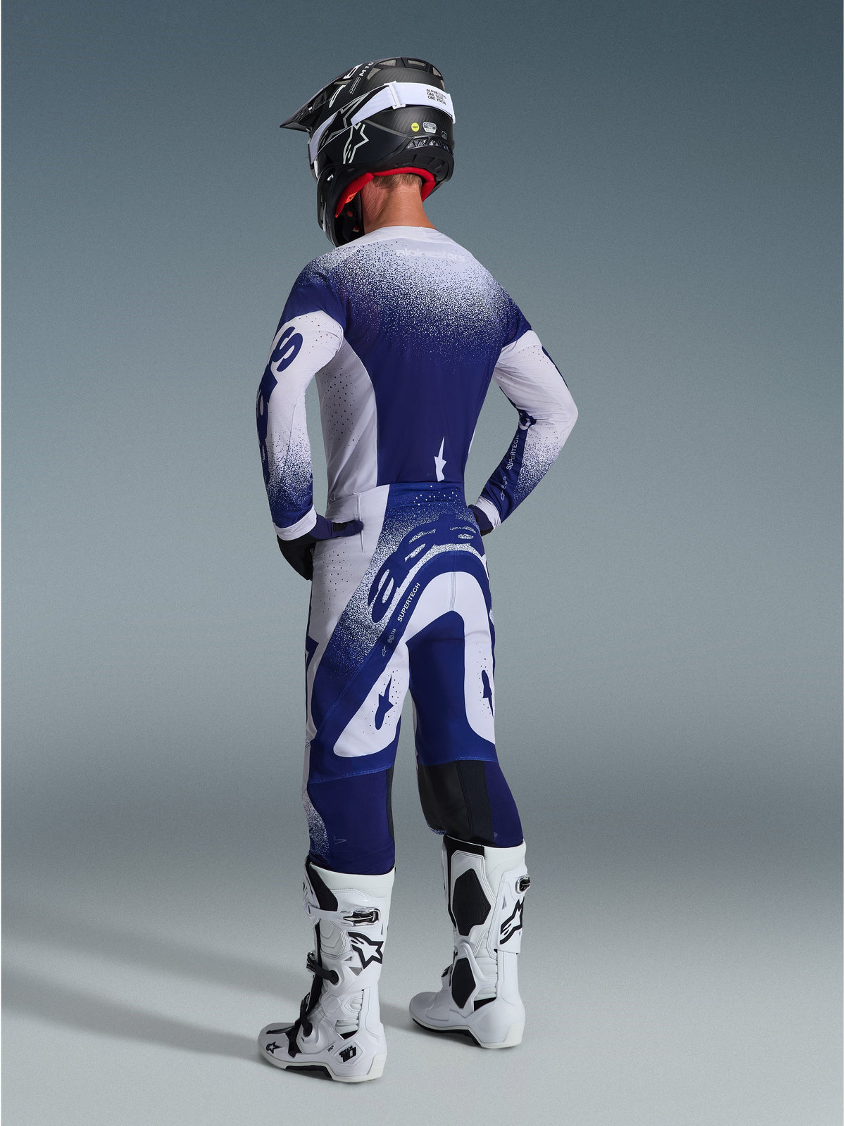 Alpinestars 2026 Supertech Scenz Pants, calças de motocross, cinzento claro e azul escuro, vista traseira com camisola a condizer e botas brancas, com grafismos de pontos em gradiente e ajuste atlético de performance para corridas todo-o-terreno