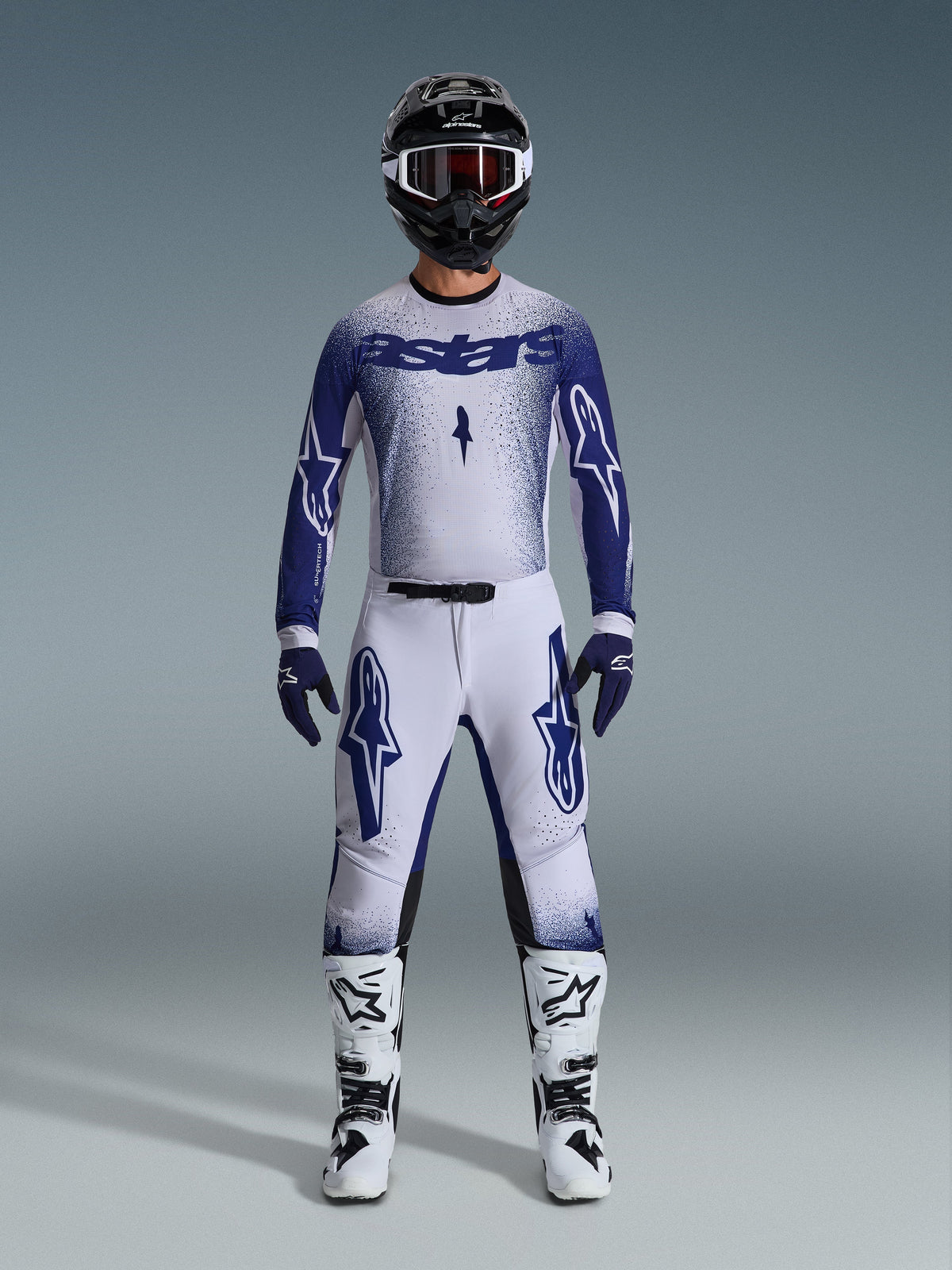 Piloto de motocross com o conjunto de equipamento Alpinestars 2026 Supertech Scenz, cinzento claro e azul escuro, incluindo camisola e calças com luvas azuis a condizer, botas brancas e pretas, e capacete preto com óculos brancos, fato coordenado para corridas profissionais de todo-o-terreno