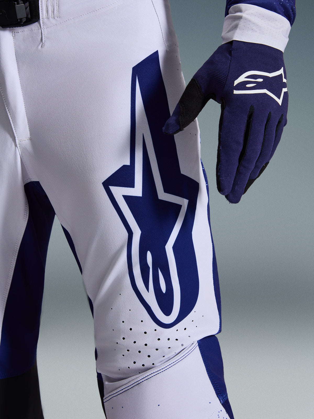 Alpinestars 2026 Supertech Scenz Pants, calças de motocross, cinzento claro e azul escuro, vista de pormenor da coxa com logótipo Alpinestars grande em azul escuro, zonas de ventilação perfuradas e tecido rip-stop elástico, usadas com uma luva de motocross azul escuro a combinar