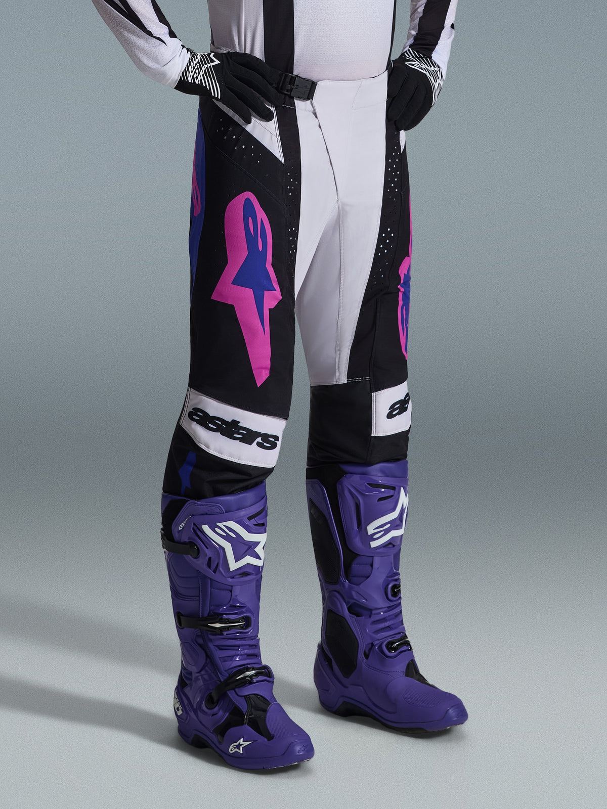Alpinestars 2026 Techstar Knif Pants, em preto, cinzento claro e roxo, usadas com botas de motocross roxas a condizer, com detalhes cor-de-rosa vibrantes e marca 'astars', equipamento profissional de motocross