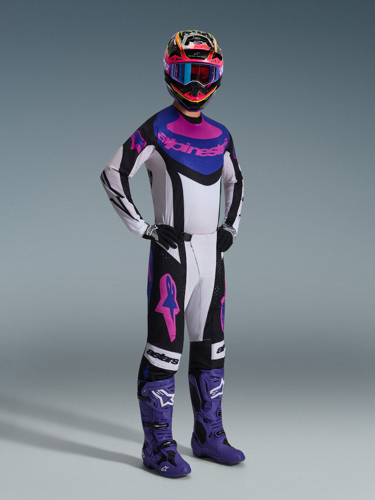 Piloto de motocross a usar o conjunto Alpinestars 2026 Techstar Knif, preto, cinzento claro e roxo, camisola branca com detalhes azuis e cor-de-rosa, calças a condizer com logótipos de estrela roxos, capacete colorido em cor-de-rosa e amarelo, botas de motocross roxas e luvas pretas, equipamento profissional para corridas de todo-o-terreno