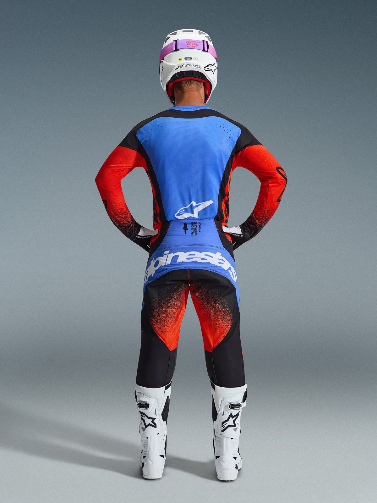 Motocross rider seen from behind wearing the 2026 Techstar Knif Pants in Orange, Black, and UCLA Blue, coordinated with a blue and orange jersey, white helmet with pink goggles, and white racing boots, full Alpinestars off-road competition gear