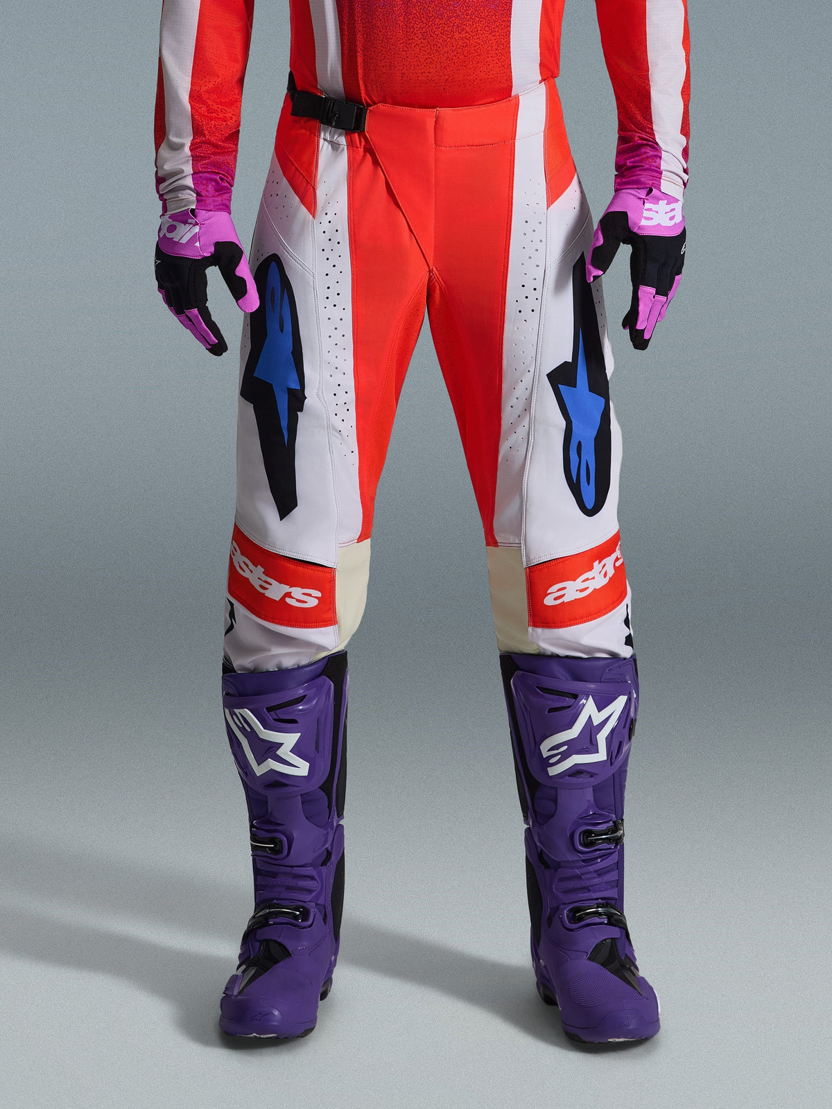 Alpinestars 2026 Techstar Knif Pants, calças de motocross em laranja fluorescente e cinzento claro com detalhes em preto, apresentadas com botas e luvas de motocross roxas, com logótipos de estrela azuis e construção de múltiplos painéis para desempenho todo-o-terreno
