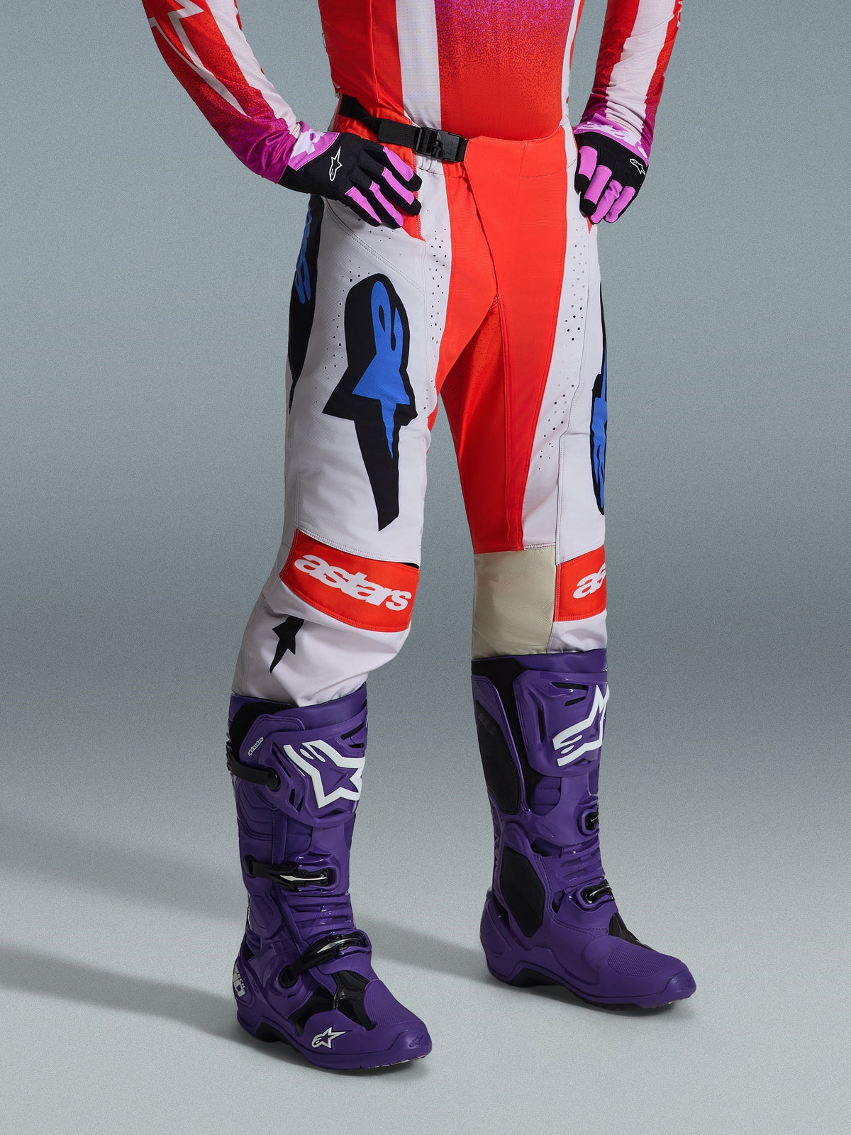 Calças Alpinestars 2026 Techstar Knif, calças de motocross, brancas e vermelho vivo com detalhes gráficos azuis, com o logótipo "astars" no joelho, usadas com botas roxas e luvas pretas e cor-de-rosa, conjunto de equipamento profissional de corrida off-road.