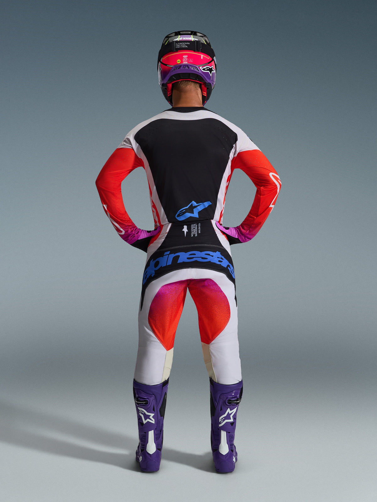 Piloto de motocross visto de costas a usar calças Techstar Knif 2026 e camisola a condizer, design em cinzento claro e preto com detalhes em laranja vibrante e roxo, logótipo Alpinestars azul no assento, botas e capacete roxos, conjunto de equipamento profissional de corrida off-road.
