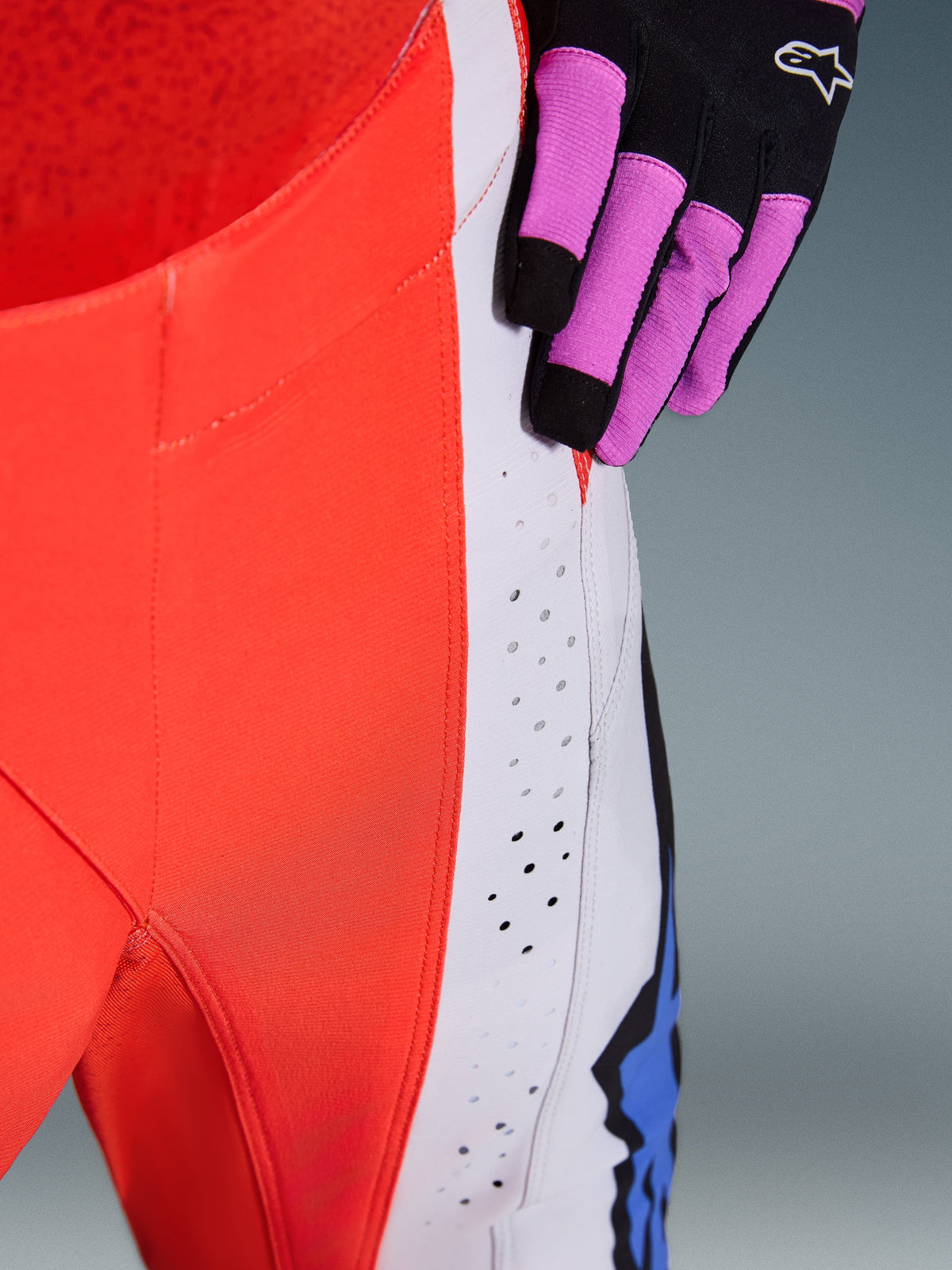Alpinestars 2026 Techstar Knif Pants, calças de motocross, em laranja fluo e cinzento claro com detalhes azuis, grande plano das perfurações de ventilação cortadas a laser na coxa, usadas com uma luva de motocross preta e roxa