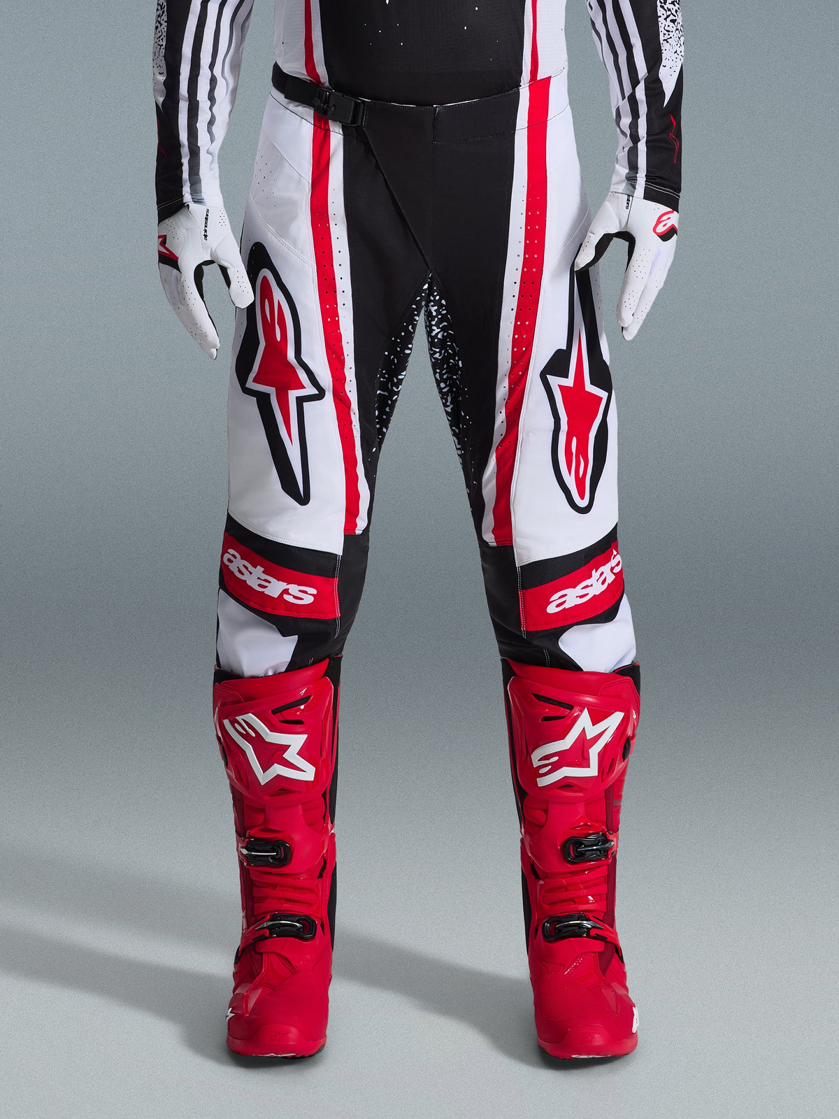 Alpinestars 2026 Techstar Nomur Pants, calças de motocross, pretas, brancas e vermelhas, usadas com botas vermelhas a condizer e luvas brancas, com logótipos vermelhos destacados nas coxas e marca 'astars' nos joelhos, design de corrida multi-painel para desempenho em todo-o-terreno