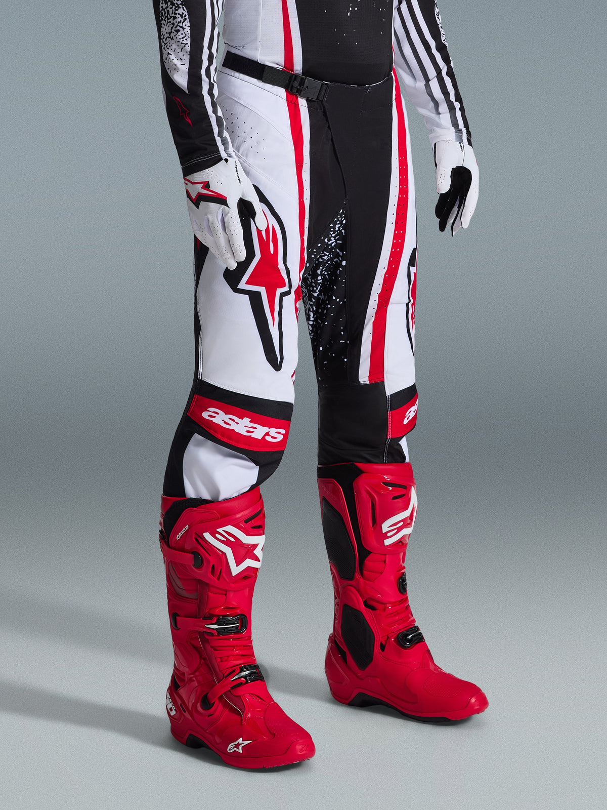 Alpinestars 2026 Techstar Nomur Pants, calças de motocross, pretas, brancas e vermelhas, usadas com botas de corrida vermelhas vibrantes e luvas brancas, apresentando riscas verticais marcantes, um logótipo de estrela vermelha grande na coxa e painéis de joelho reforçados para performance em todo-o-terreno