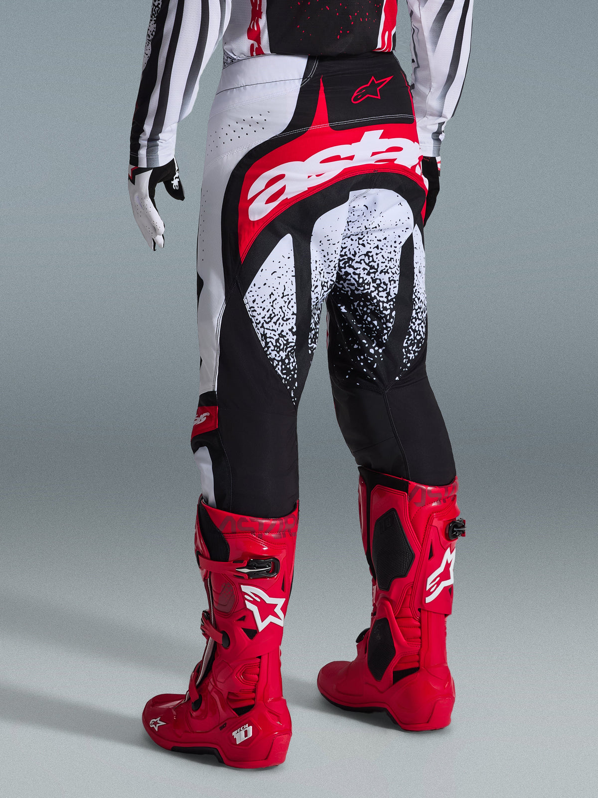 Alpinestars 2026 Calças Techstar Nomur, calças de motocross, pretas, brancas e vermelhas, vista traseira num piloto com botas vermelhas a condizer, com logótipo 'astars' proeminente em vermelho e branco no assento e padrões gráficos salpicados, equipamento de corrida profissional para todo-o-terreno