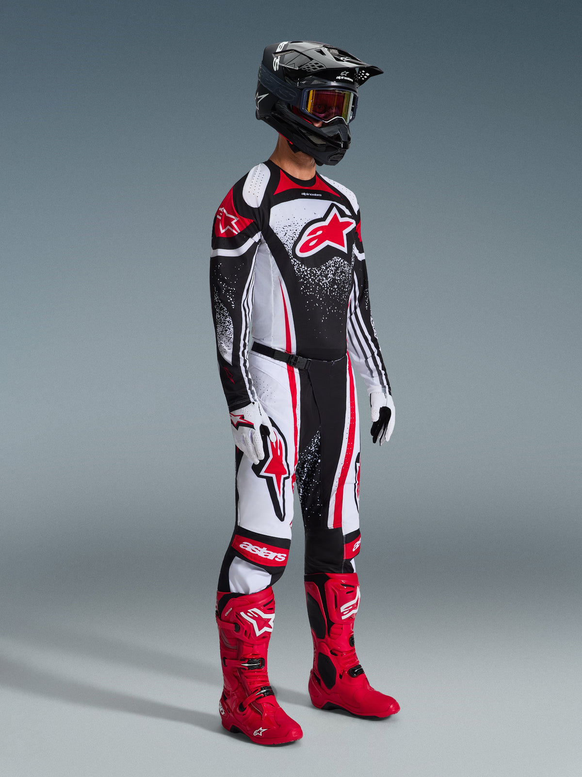 Piloto de motocross com equipamento de corrida completo, calças Techstar Nomur 2026 em preto, branco e vermelho, camisola a condizer com logótipo Alpinestars proeminente e design salpicado, botas de motocross em vermelho vivo, luvas brancas e pretas, e capacete preto com óculos de proteção, conjunto profissional de corrida todo-o-terreno coordenado.