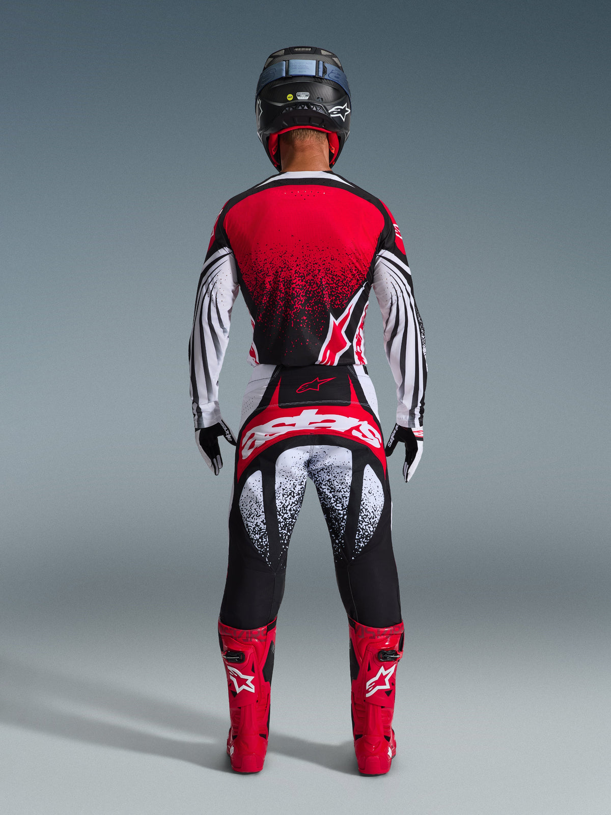 Vista traseira de um piloto de motocross a usar calças Alpinestars 2026 Techstar Nomur em Preto Branco Vermelho, apresentando um logótipo em destaque no assento e grafismos salpicados, em conjunto com uma camisola vermelha e botas vermelhas vibrantes para um equipamento de competição todo-o-terreno completo