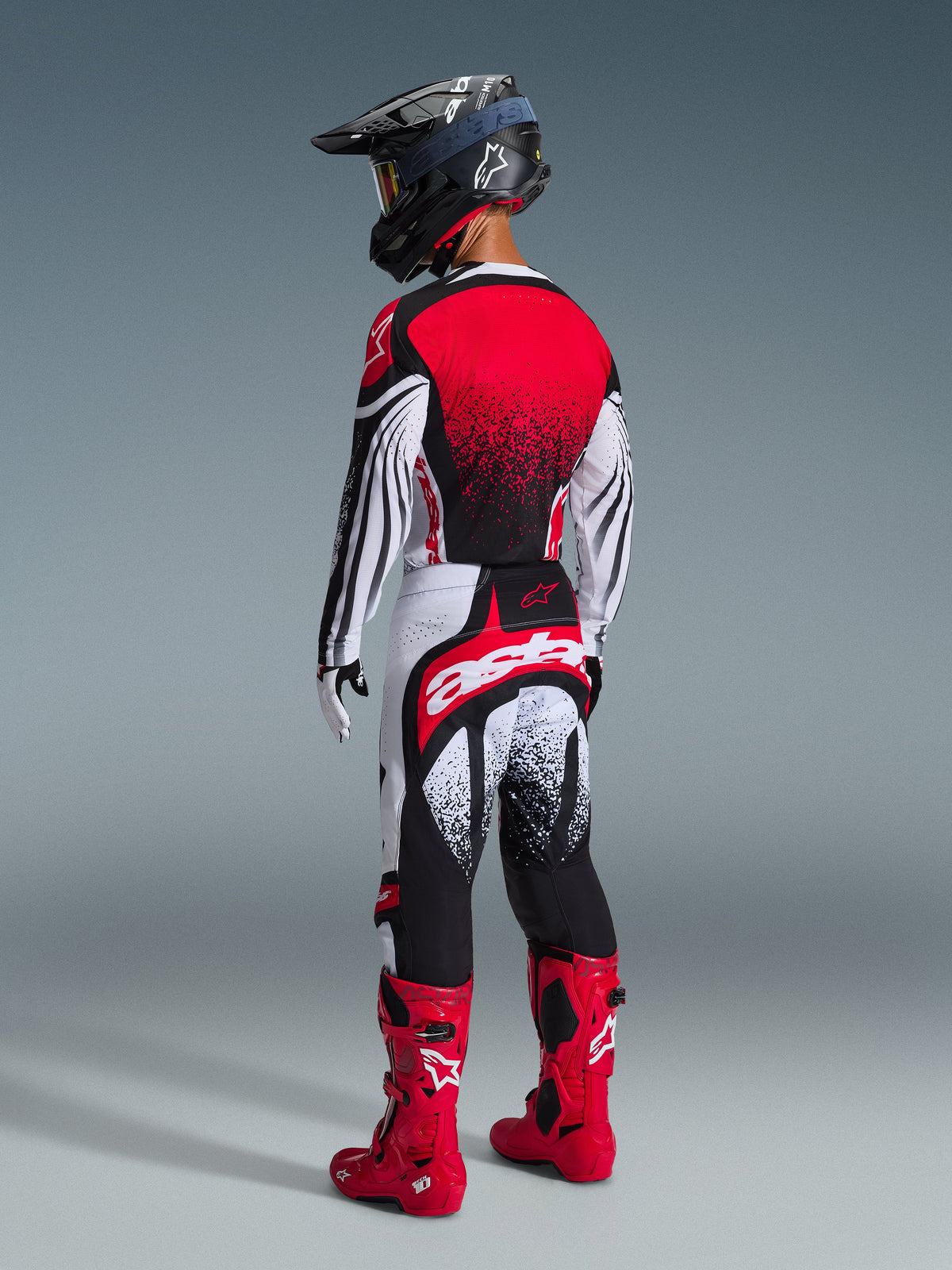 Piloto de motocross visto de costas a usar as calças Alpinestars 2026 Techstar Nomur em preto, branco e vermelho, com um grande logótipo 'astars' branco no assento, combinadas com uma camisola a condizer, botas de motocross em vermelho vivo e um capacete preto, conjunto de equipamento profissional para corridas todo-o-terreno