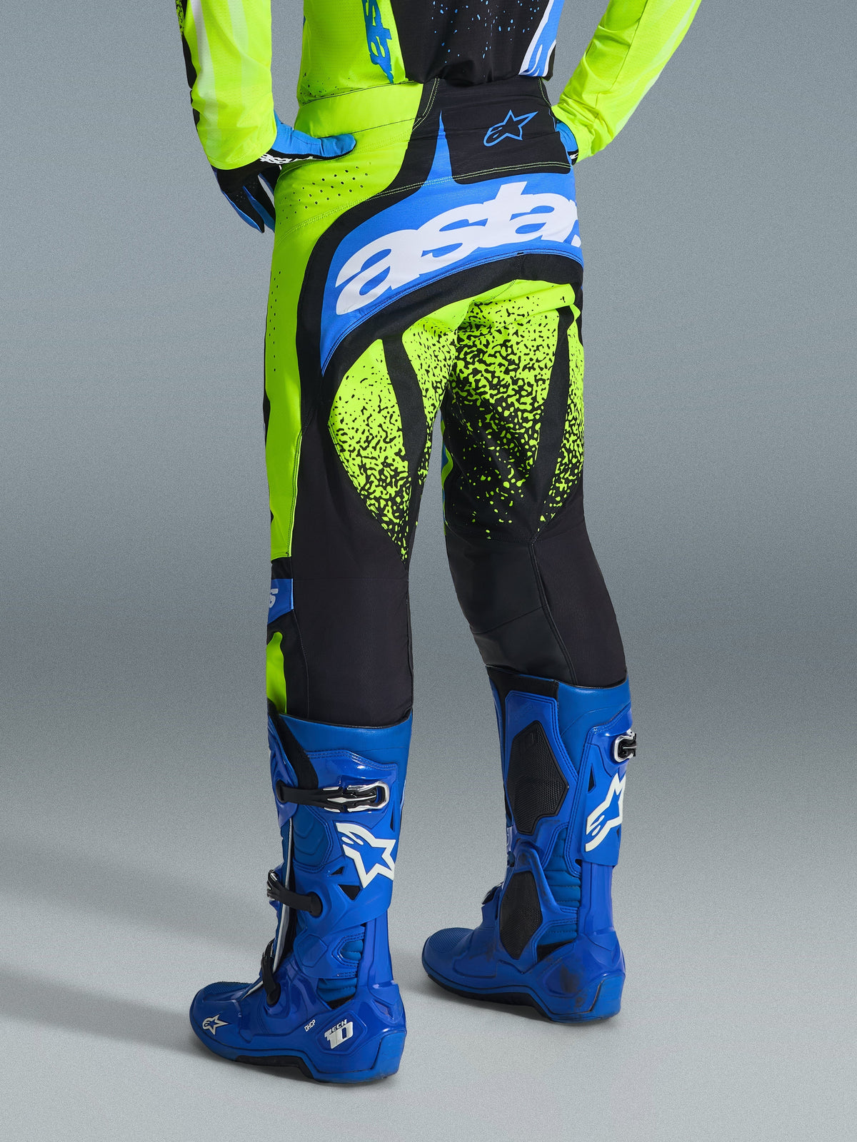 Alpinestars 2026 Techstar Nomur Pants, calças de motocross, Amarelo Fluo e Azul UCLA, vista traseira do piloto a usar botas azuis, com a marca 'astars' no assento e grafismos de padrão de salpicos pretos nas pernas