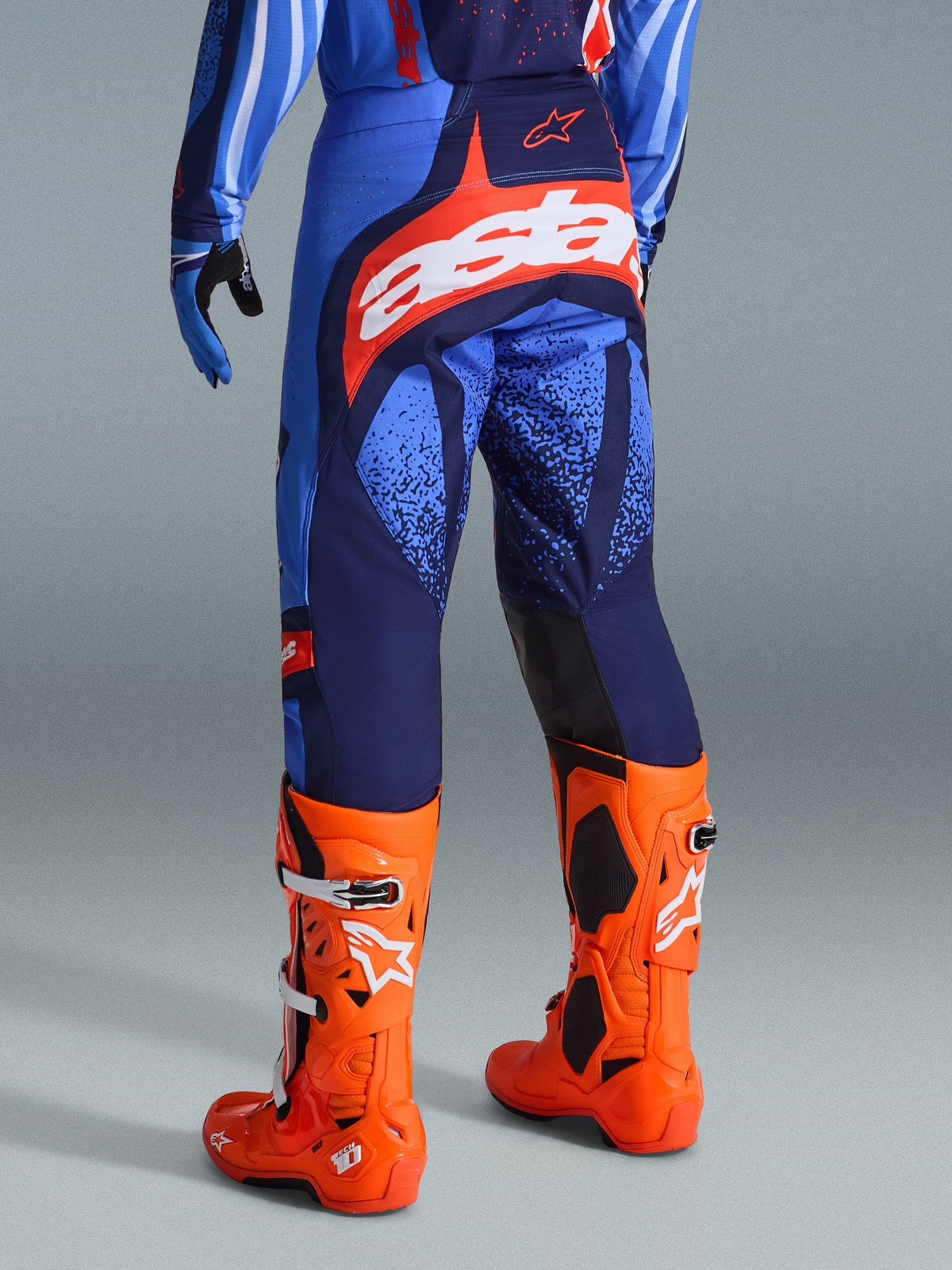 Alpinestars 2026 Techstar Nomur Pants, calças de motocross, azul-escuro, laranja e azul UCLA, vista traseira com logótipo 'Astars' branco no assento, usadas com botas de motocross cor de laranja vibrante e camisola a condizer, corte atlético de corrida para competição fora de estrada