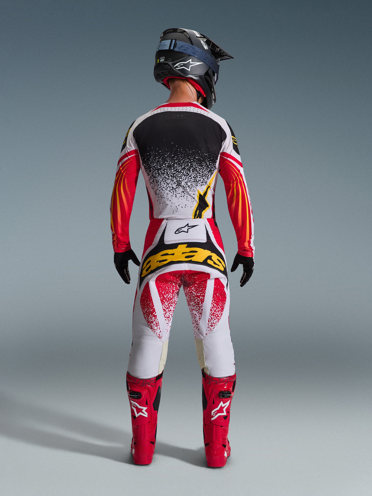 Conjunto de equipamento Alpinestars 2026 Techstar Nomur, equipamento completo de motocross visto de costas, camisola branca com efeito de salpicos pretos e mangas com grafismos vermelho e laranja, calças brancas e vermelhas com logótipo 'astars' amarelo, botas de corrida vermelhas, capacete preto com textura de carbono e óculos azuis, equipamento de alto desempenho para todo-o-terreno