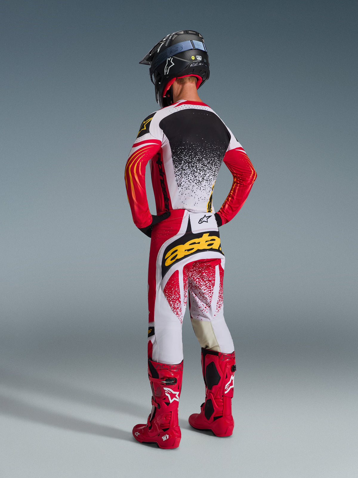 Piloto de motocross visto de costas com o Conjunto de Equipamento Alpinestars 2026 Techstar Nomur, camisola vermelha, branca e preta com padrão salpicado em branco nas costas, calças a condizer com o logótipo 'astars' amarelo no assento, botas de corrida em vermelho vivo e capacete preto, equipamento de motocross completo e coordenado