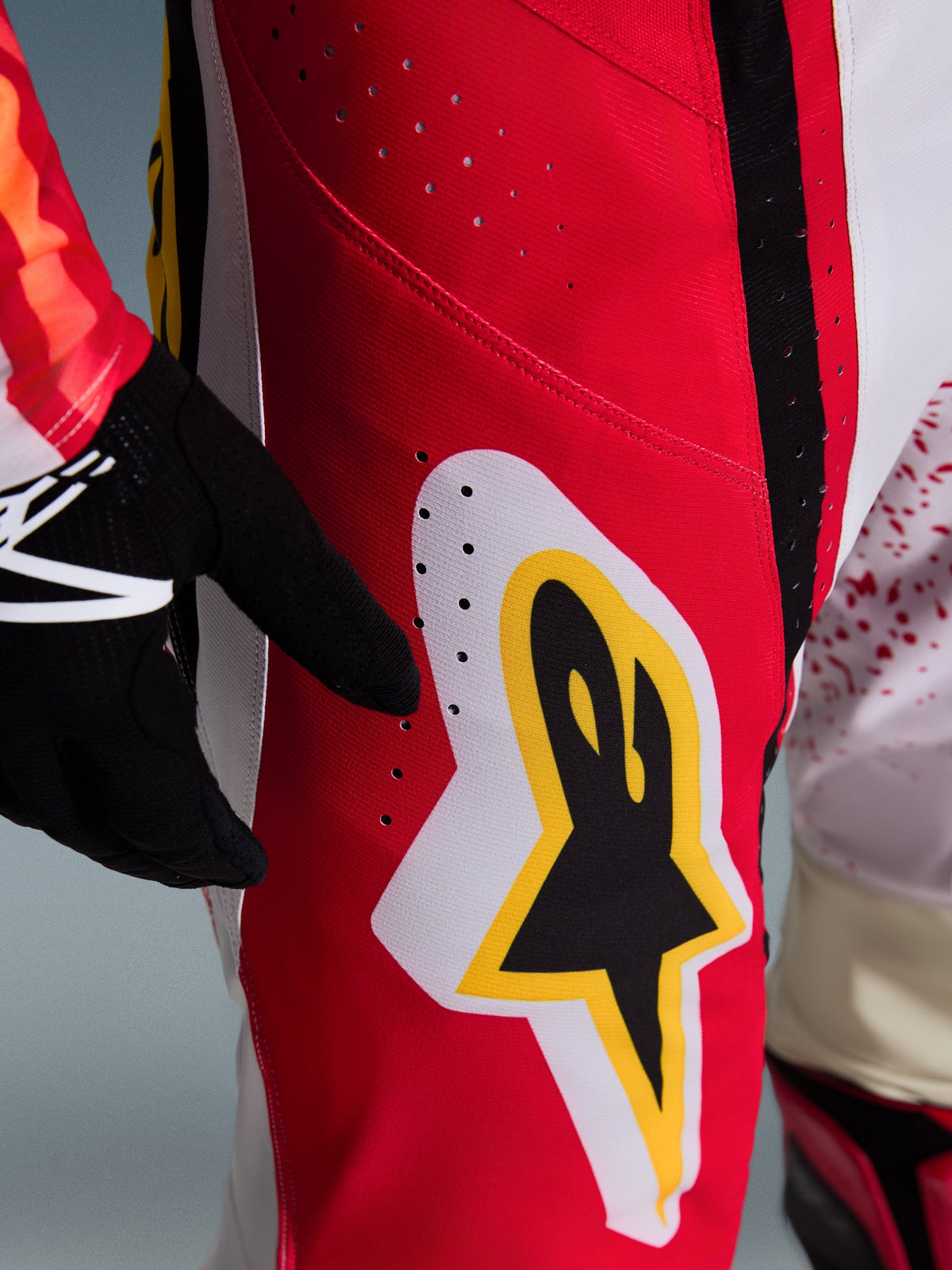 Calças Alpinestars Techstar Nomur 2026, detalhe dos painéis vermelhos e brancos na coxa, com logótipo Astar grande em preto, amarelo e branco, perfurações a laser para ventilação, parte de um conjunto de motocross profissional