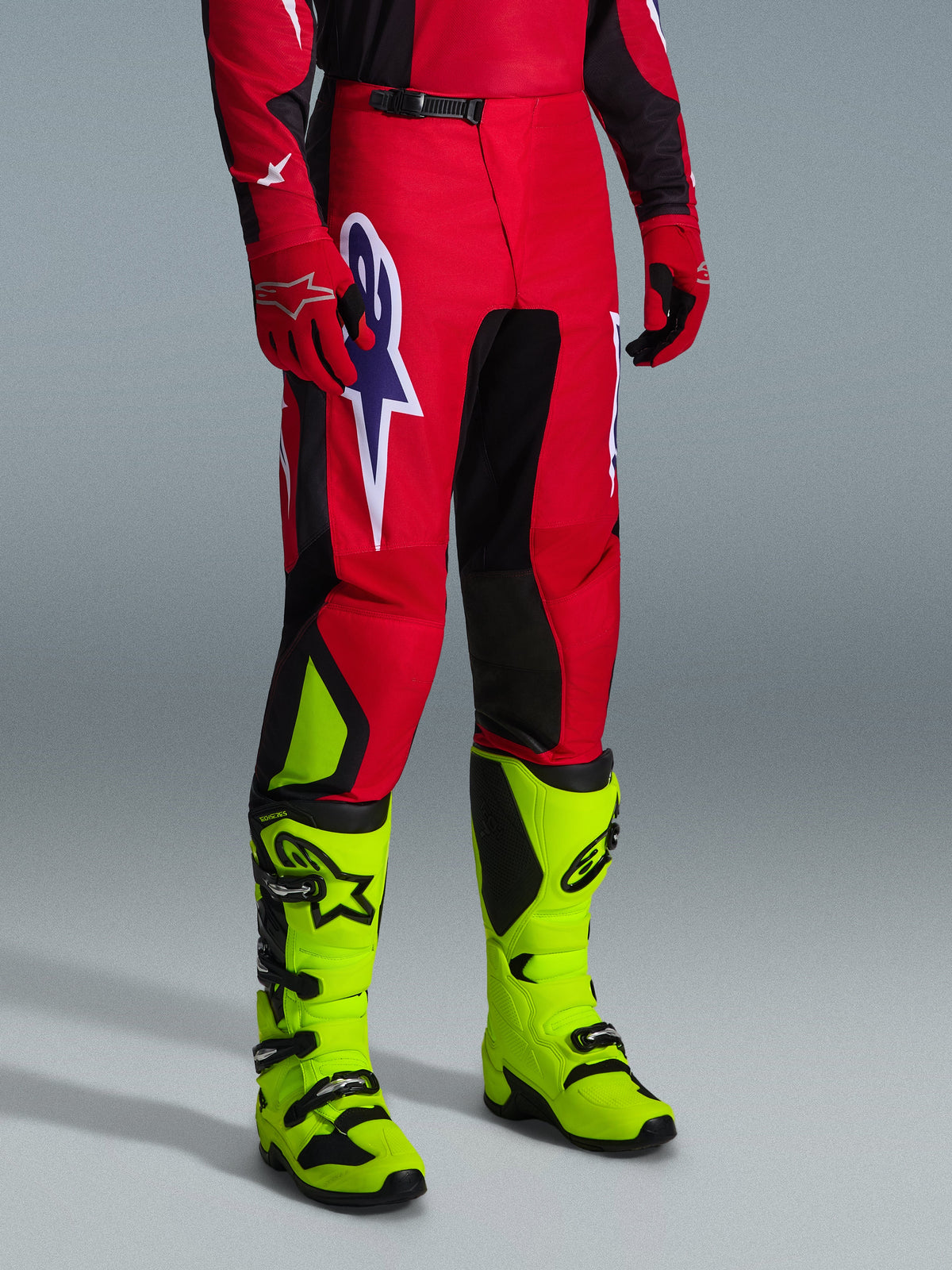 Alpinestars 2026 Fluid Grid Pants, calças de motocross, em vermelho, preto e roxo, usadas com botas amarelo néon e luvas vermelhas, com painéis pretos e um logótipo de estrela grande em roxo e branco, design durável de ajuste desportivo para corridas off-road