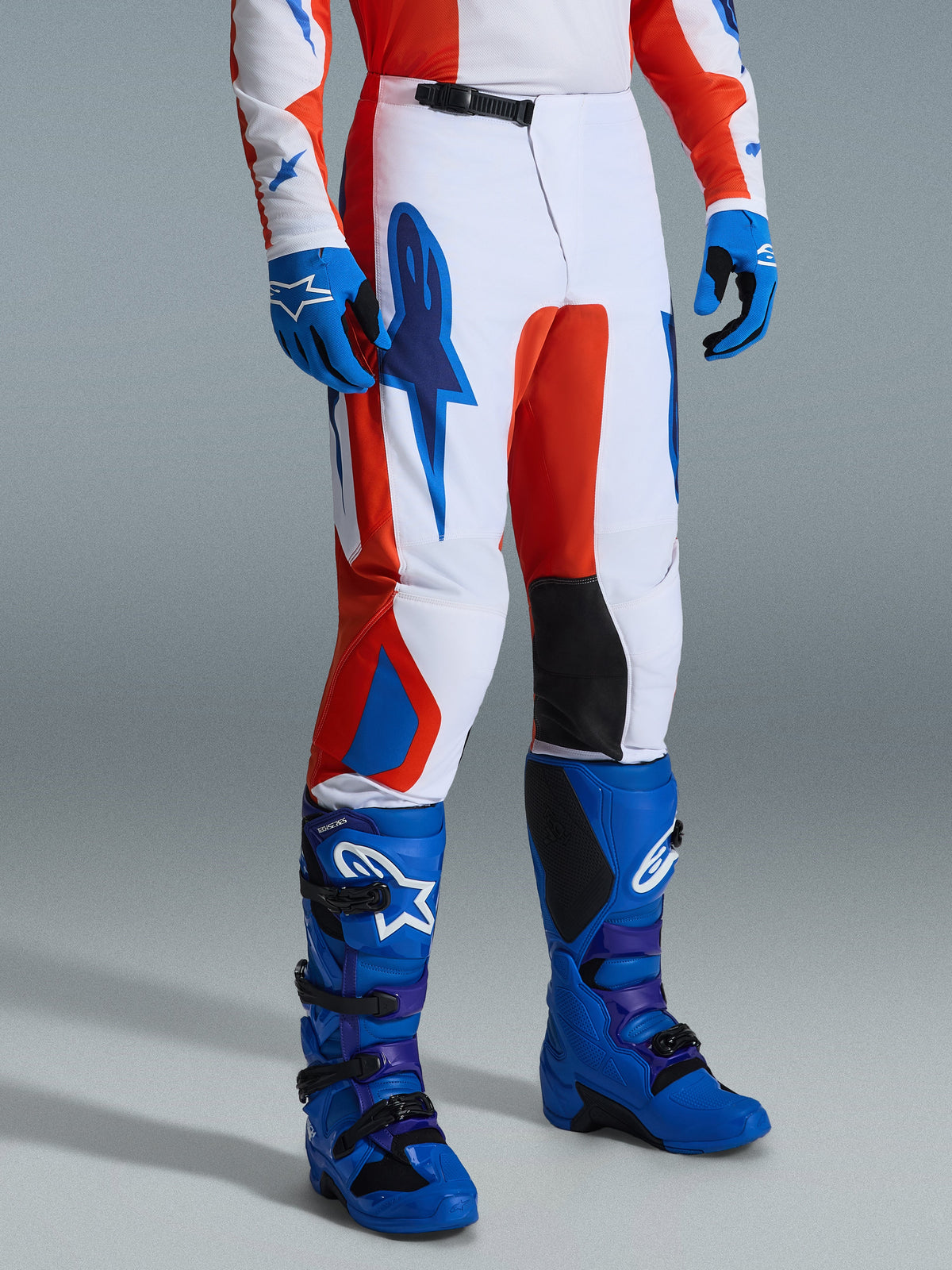 Alpinestars 2026 Fluid Grid Pants, calças de motocross, laranja e azul UCLA, usadas com luvas e botas azuis a condizer, com uma base branca e grafismos marcantes, painéis internos pretos nos joelhos e um design ergonómico para condução off-road.