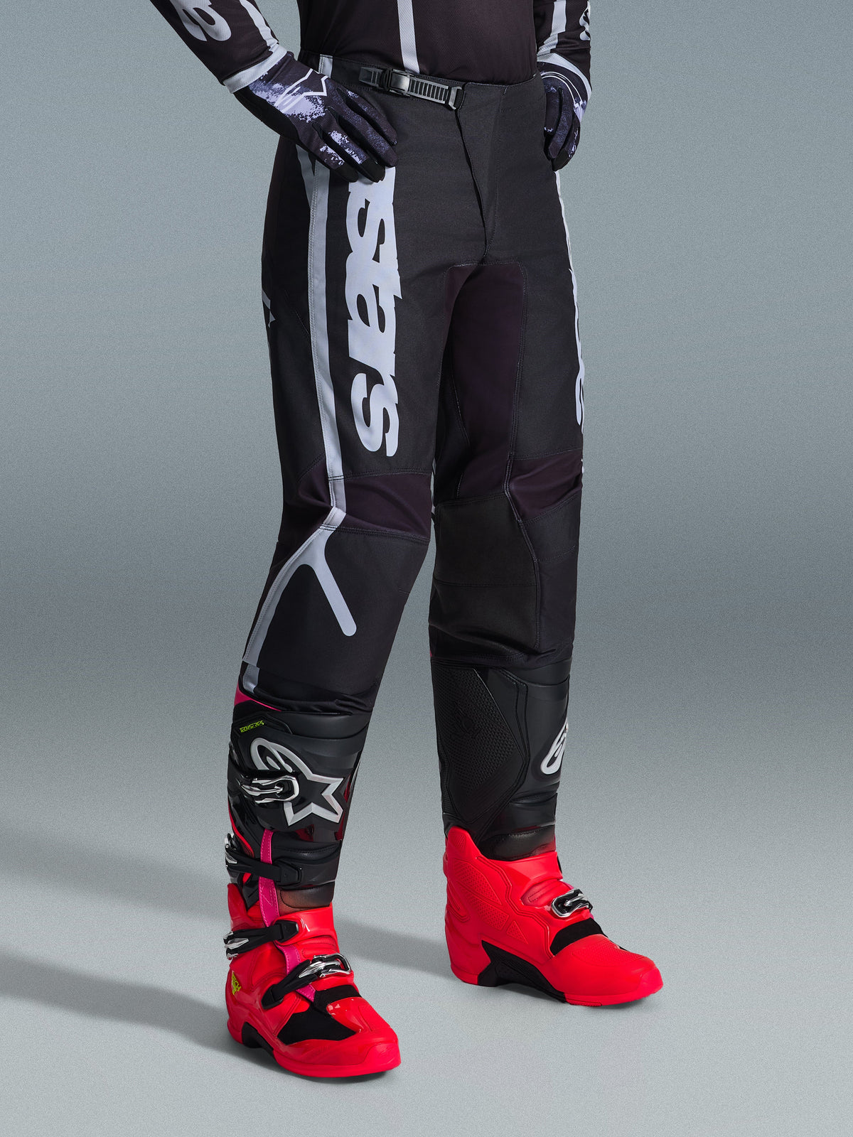 Calças Alpinestars 2026 Fluid Apex, calças de motocross, pretas e cinzentas, usadas com botas rosa vibrante e vermelhas e camisola preta e branca a combinar, com a marca Alpinestars vertical e arrojada na coxa, painéis reforçados e fecho de catraca na cintura para desempenho todo-o-terreno.