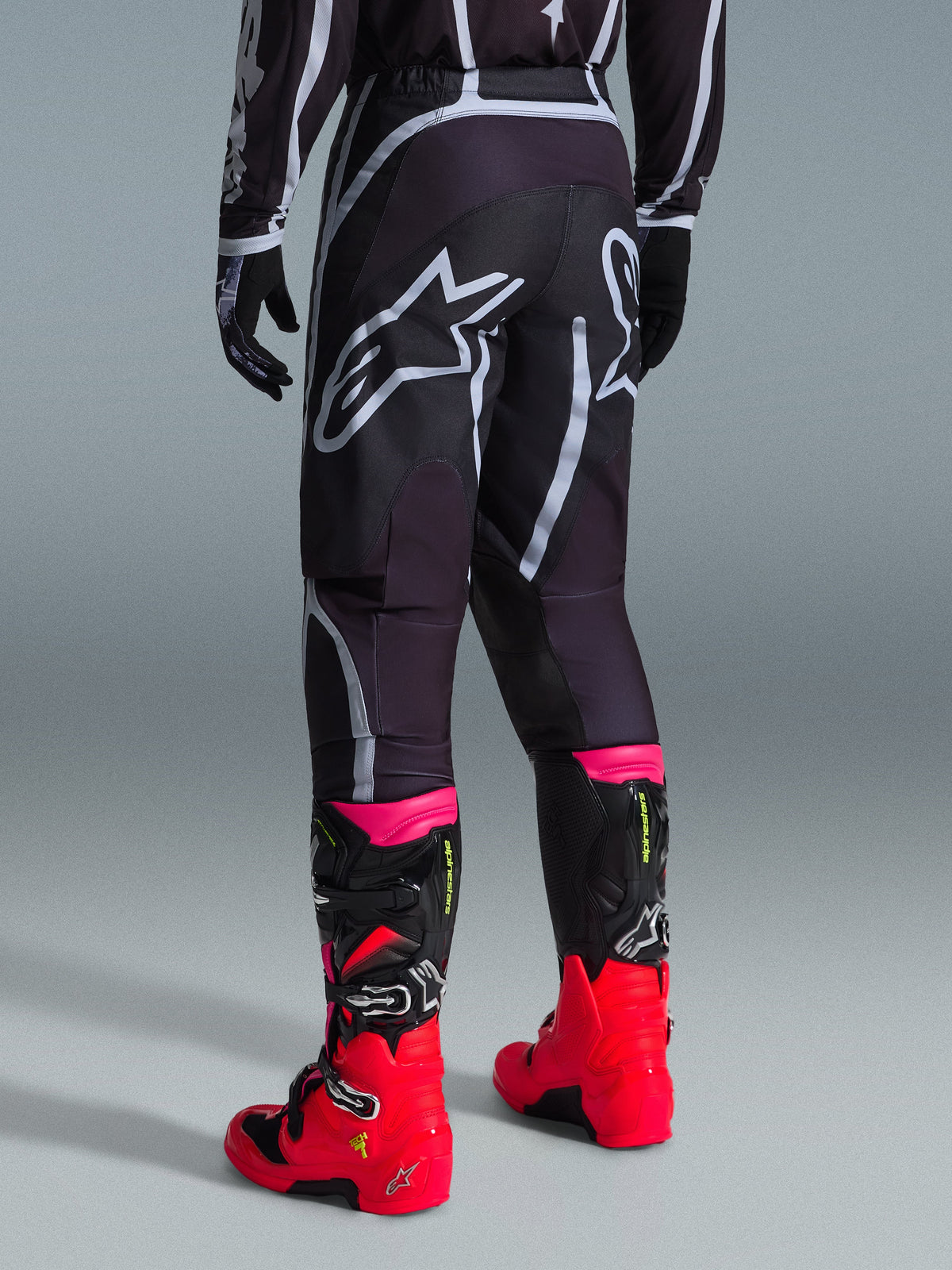 Alpinestars 2026 Fluid Apex Pants, Motocross Pants, Black and Gray, rear view featuring large white Alpinestars logos on the seat, worn with bright red and pink boots, durable 600D polyfabric construction for off-road performance